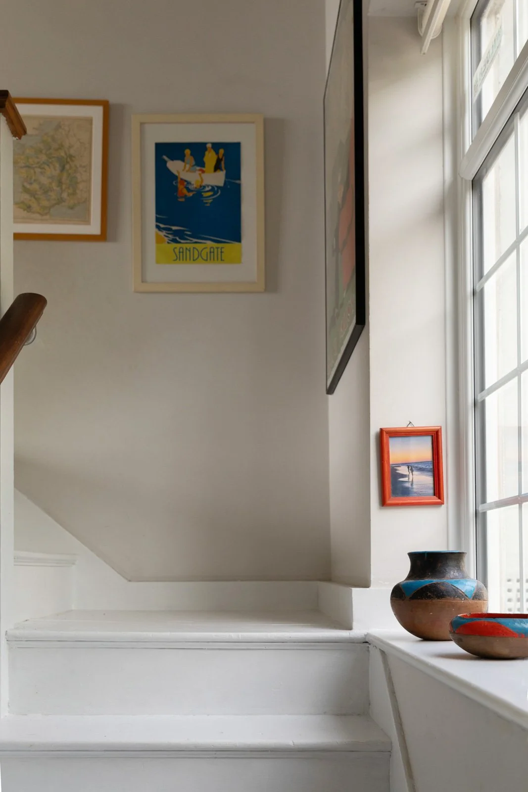 Interior view of a staircase landing with framed artwork on the walls, a large window, and pottery decorations.