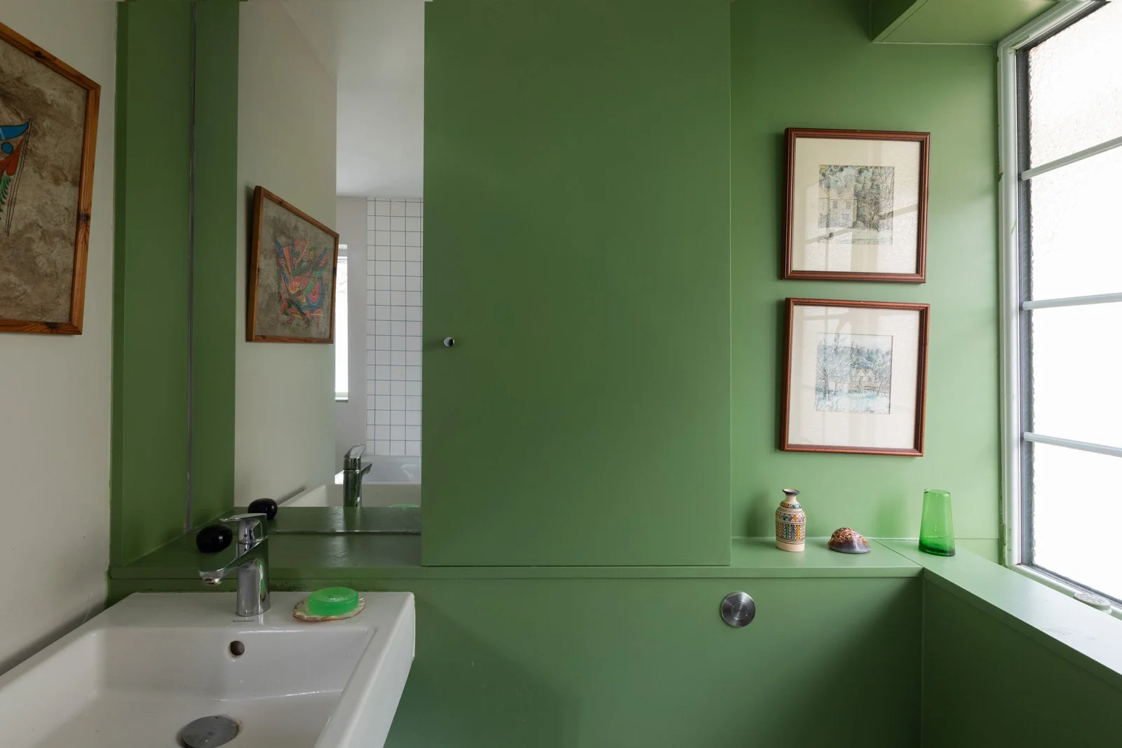 A bathroom with green walls and a small sink with a green soap on top. There is a mirror on the wall above the sink, reflecting a partial view of the room. Decorative framed pictures hang on the walls, one next to the mirror and two on the window wal
