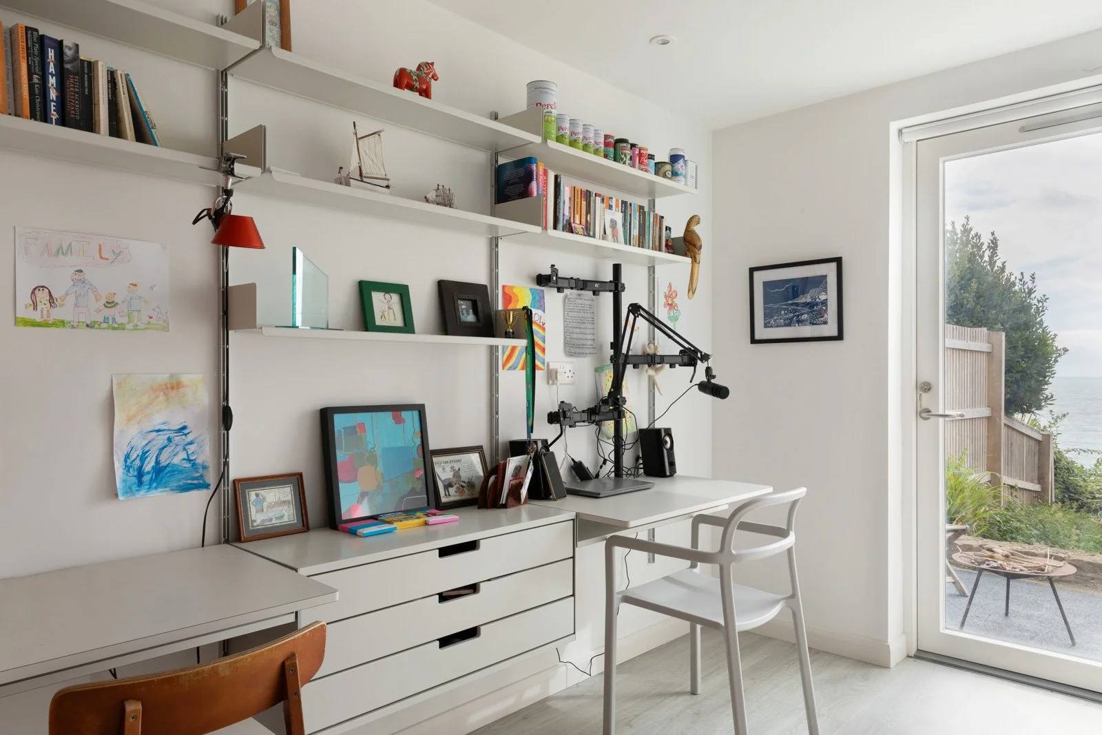 A bright home office with white furniture and shelves, featuring a desk with a microphone setup, framed pictures, books, and colorful children's drawings on the wall, with a glass door leading outside to a patio and waterfront view.