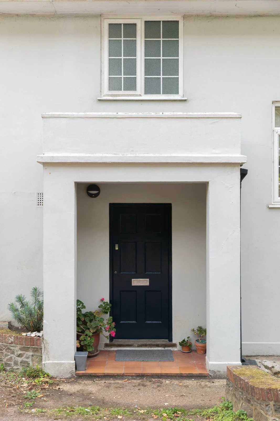 The front entrance of a white, two-story house with a black door, small plants in pots, and a tiled porch