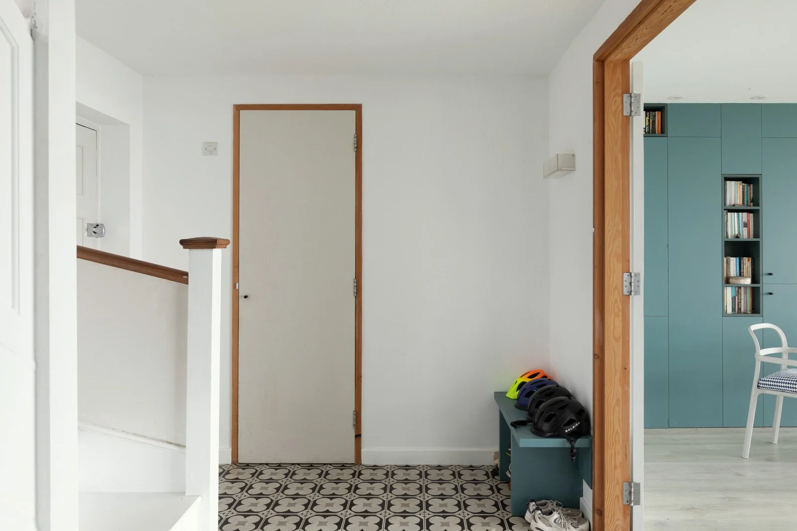 Interior view of a foyer with a white wall, closed door, and patterned tile floor, with helmets on a blue bench and a partial view of a blue bookshelf in a neighboring room.