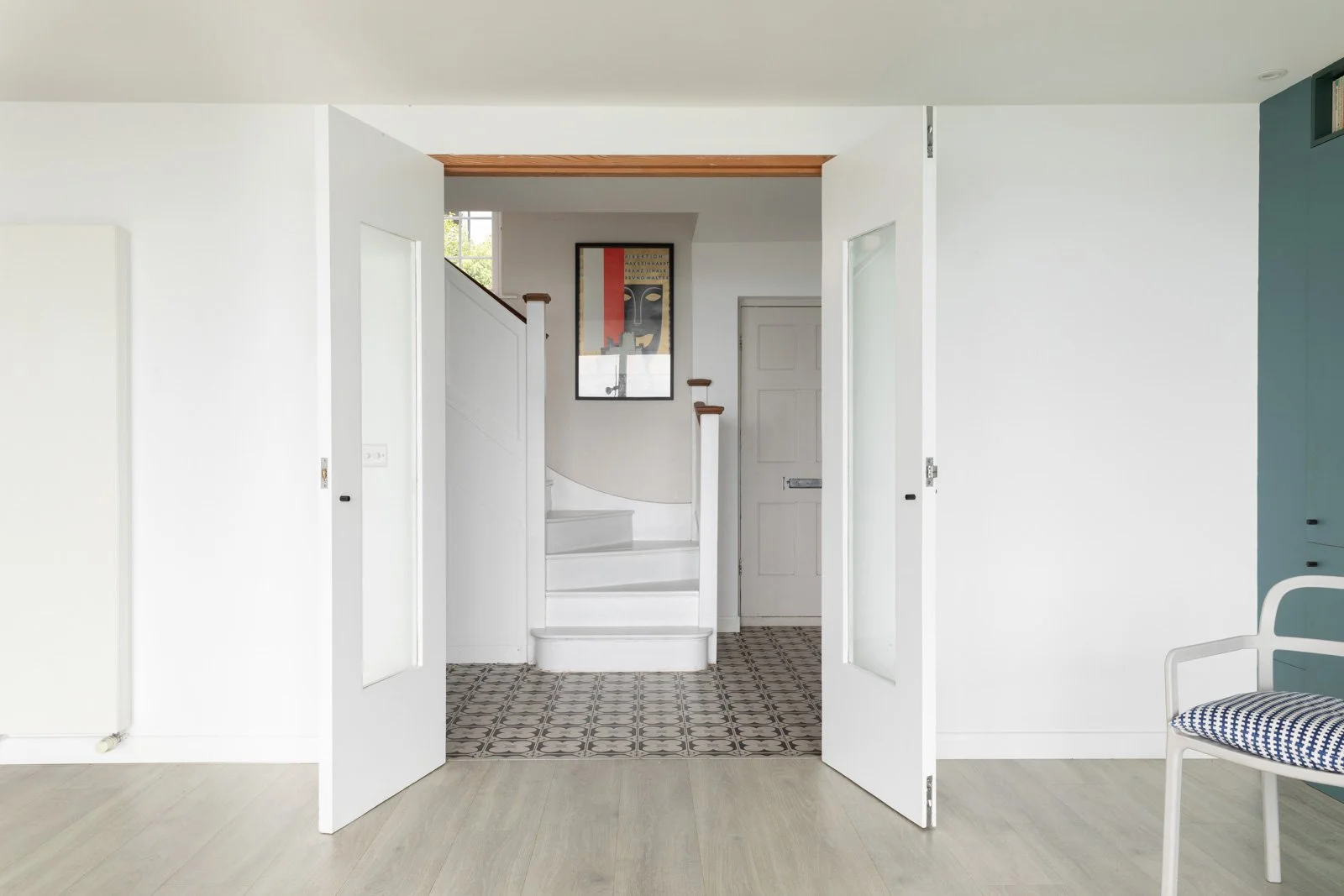 View of an interior entryway with white walls, a staircase, and a patterned tile floor, seen through open white double doors in a modern home.