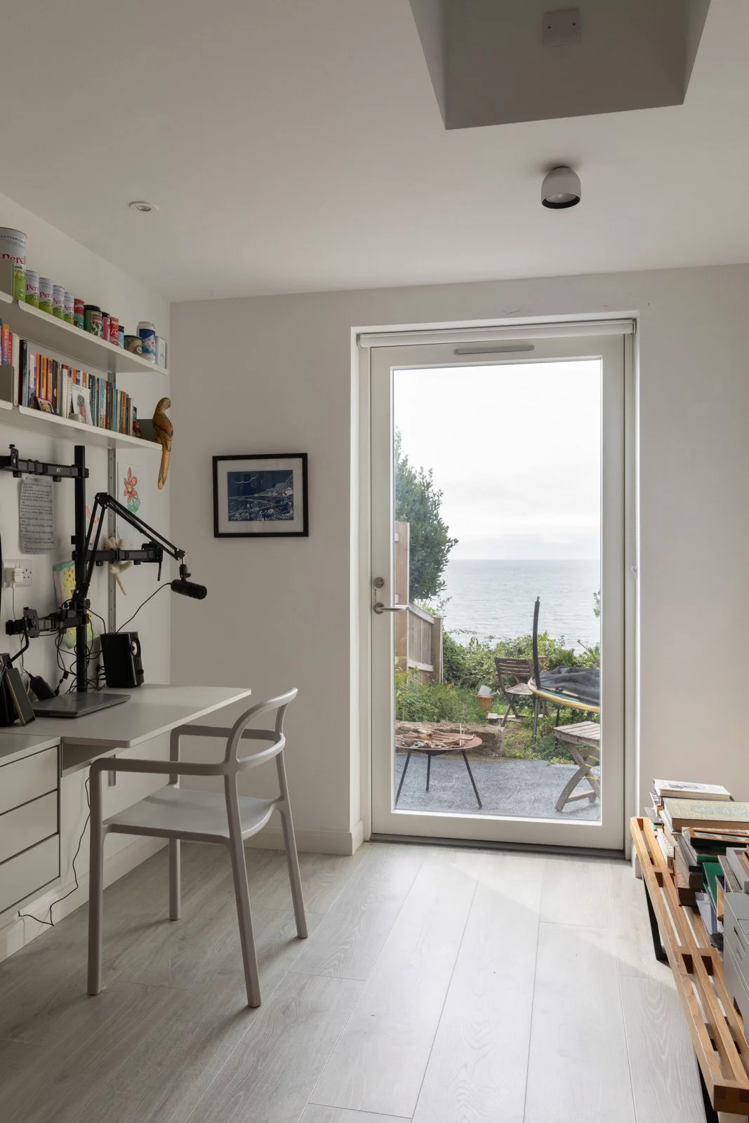 Interior view of a room with a white desk and chair, a bookshelf, a wall art, and a glass door leading to a patio with outdoor chairs and a view of the ocean.