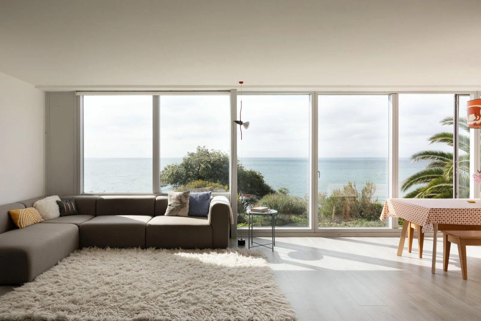 Living room with large windows overlooking the sea, gray sectional sofa with pillows, shag rug, small side table, and dining table with patterned tablecloth on the right.