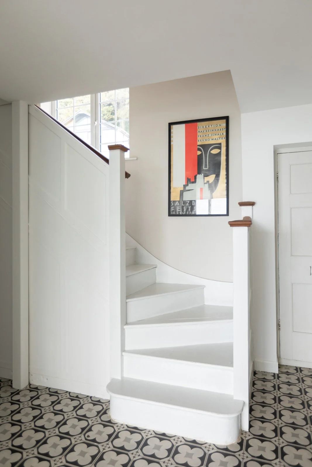 Interior view of a staircase in a house with a black and white patterned floor, white walls, a framed poster on the wall, and a window at the top of the stairs letting in natural light.