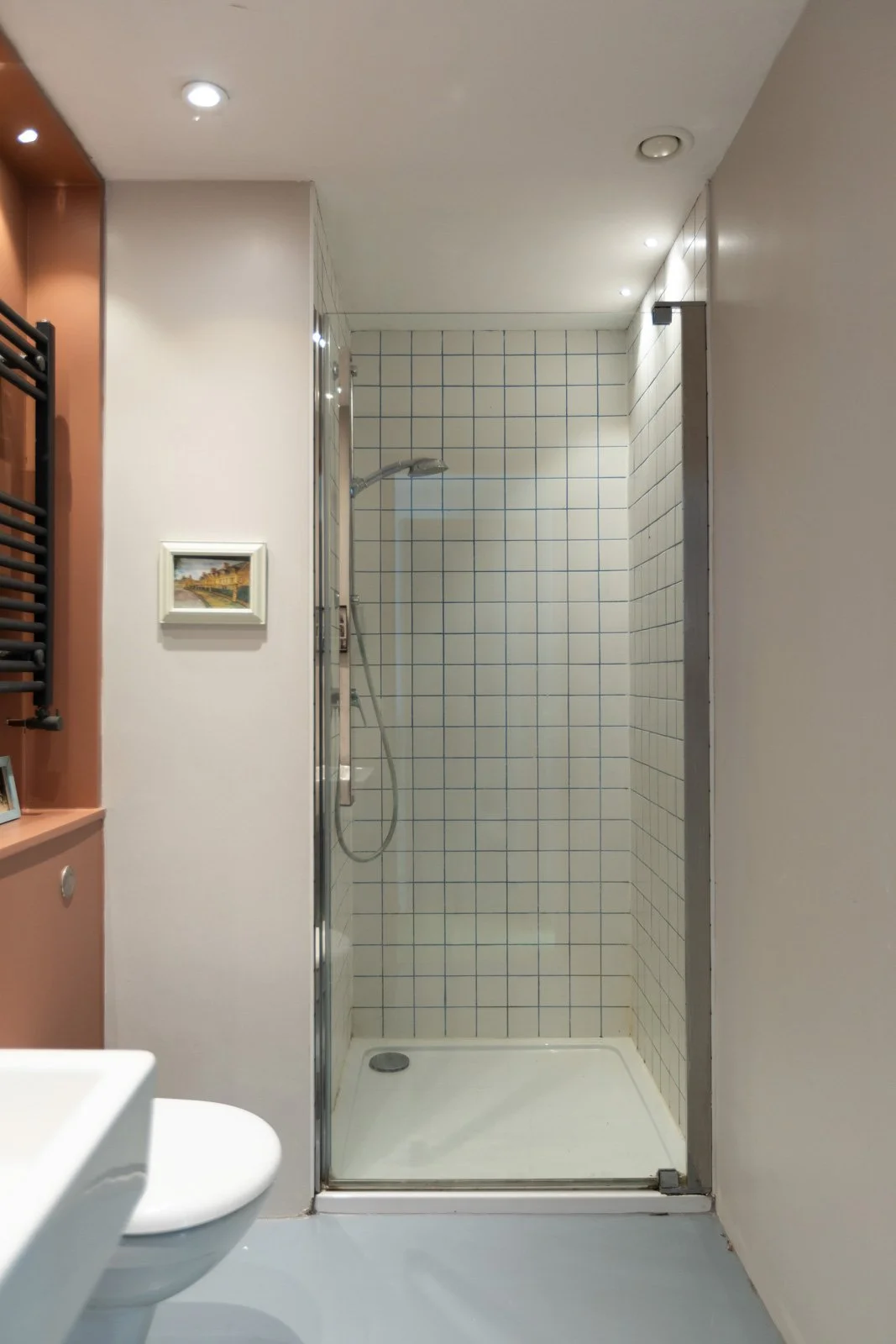Small bathroom with a walk-in shower with white tiled walls and glass sliding door, white toilet in the foreground, small artwork on the wall, and a built-in shelf with a towel rack.