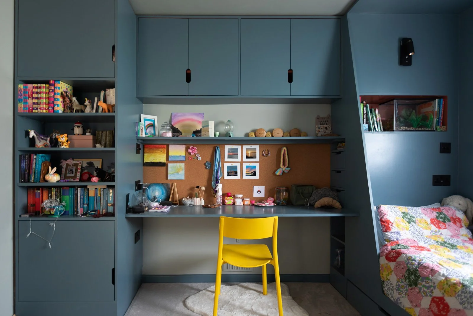 A blue built-in room with shelves, cabinets, and a study area. The left side has books, toys, and decorations. The middle has a desk with small pictures, jars, and stationery, and a yellow chair. The right side shows a bed with a colorful quilt, a st