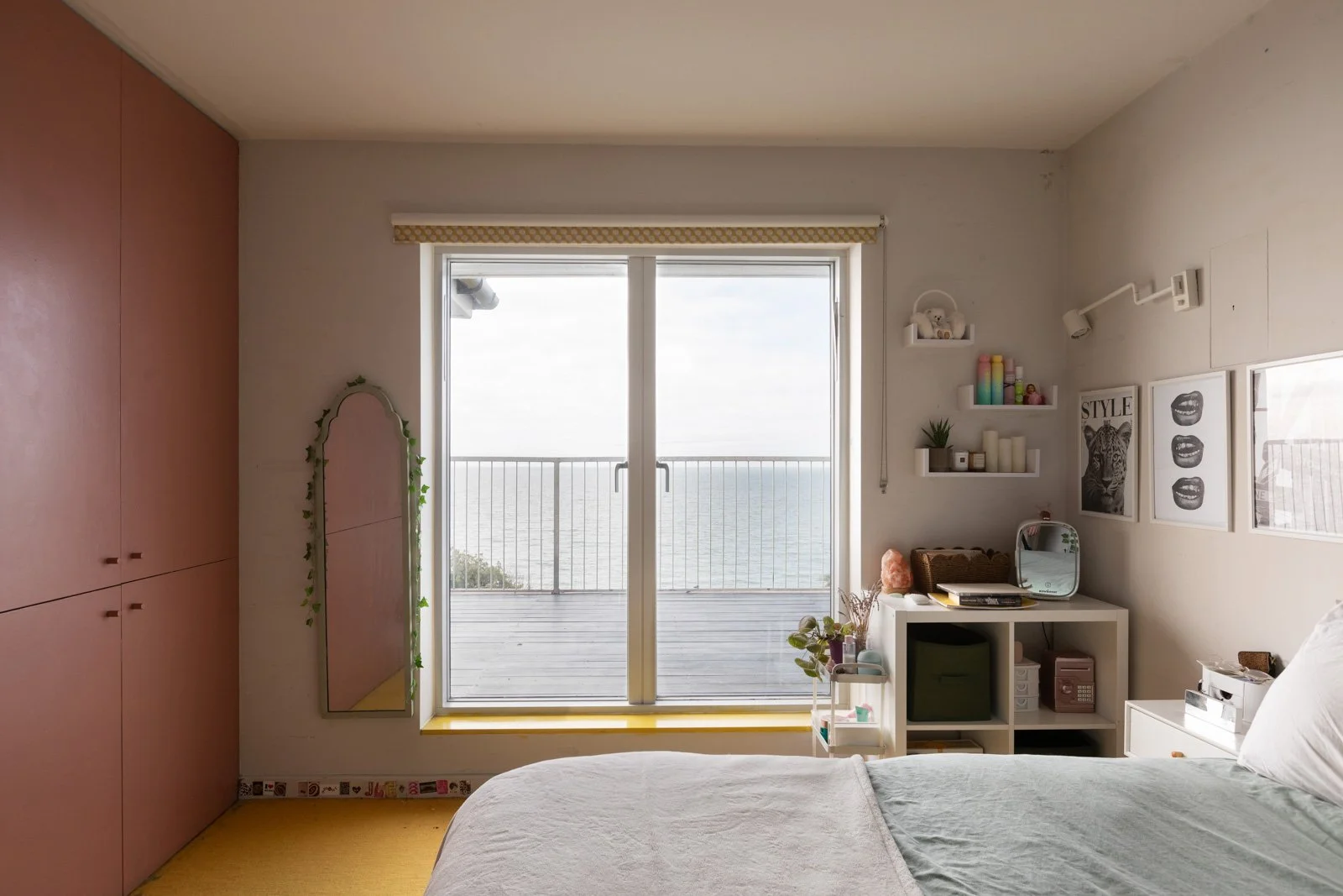 A bedroom with a large glass sliding door opening to a balcony overlooking the water. The room has pink closet doors, a mirror with a plant vine, and a white bedside table with décor. There are shelves with books and decorative items, framed artwork 