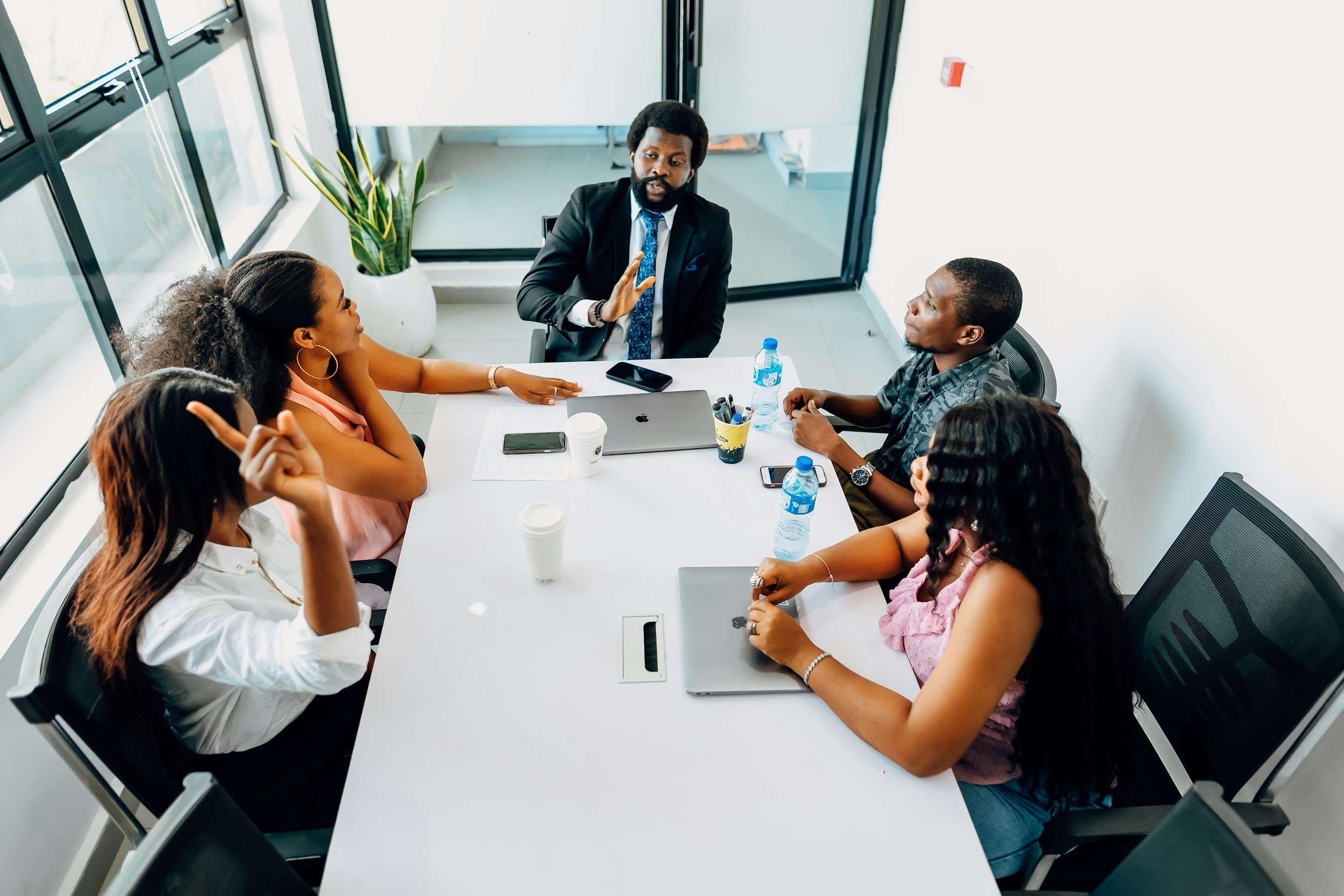 A diverse group of six people in business attire having a discussion around a conference table in a modern office. There are laptops, water bottles, and coffee cups on the table, with large windows and a plant in the background.