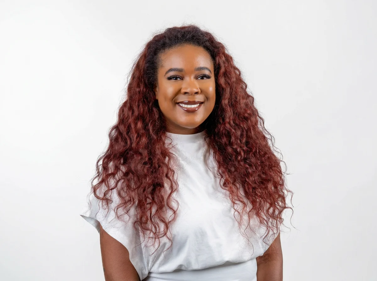 A woman with long, curly red hair smiling, wearing a white T-shirt, standing against a plain white background.