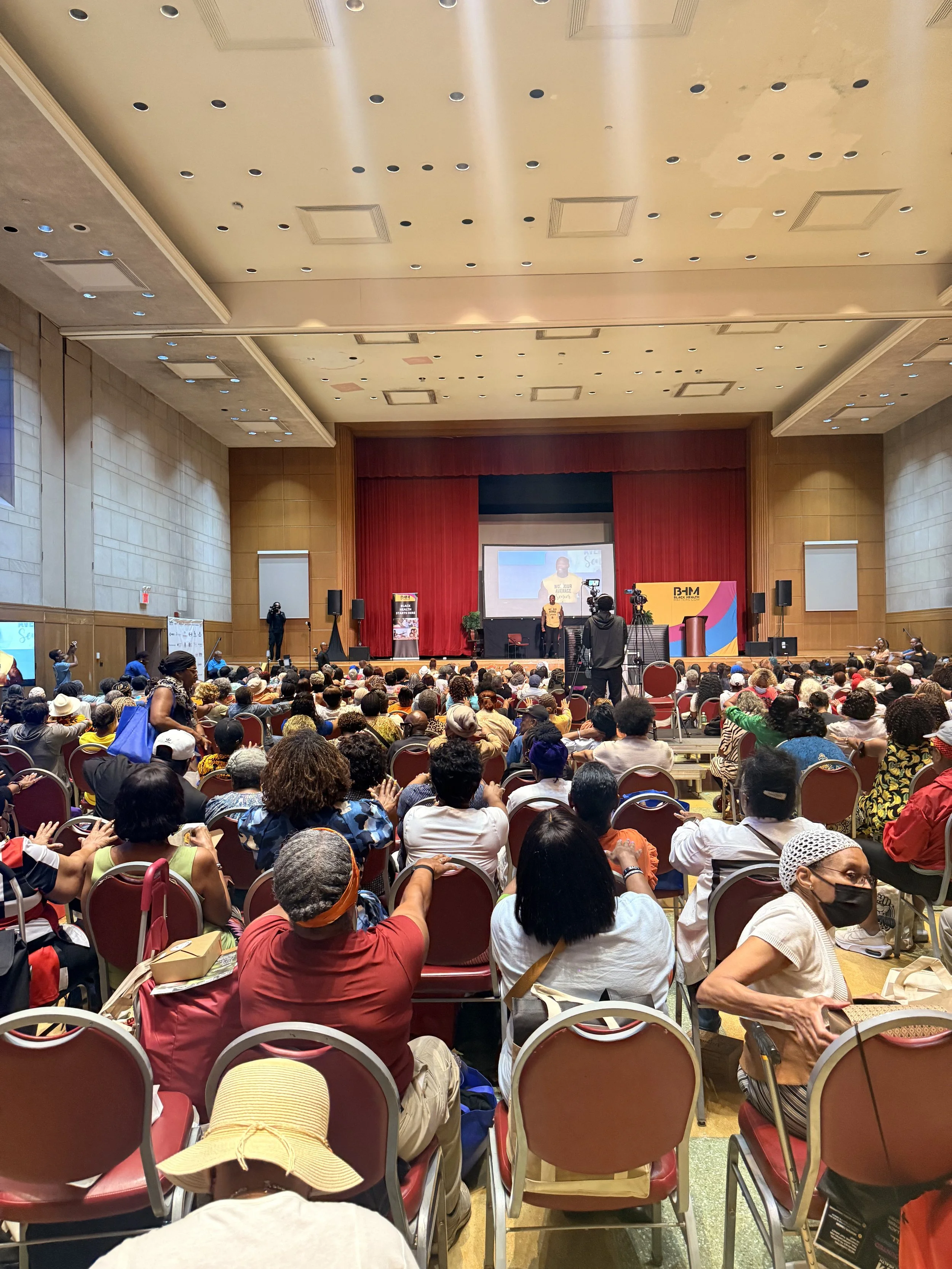 A large indoor conference or assembly with many seated attendees facing a stage with a large screen, red curtains, and speakers. The audience is diverse, wearing casual and head coverings, with some taking photos or videos.