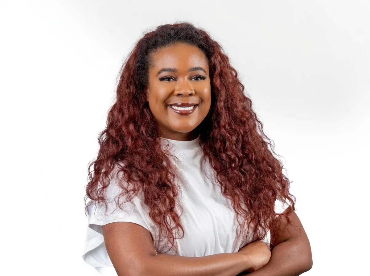 A woman with long, curly auburn hair smiling at the camera, wearing a white t-shirt against a plain white background.