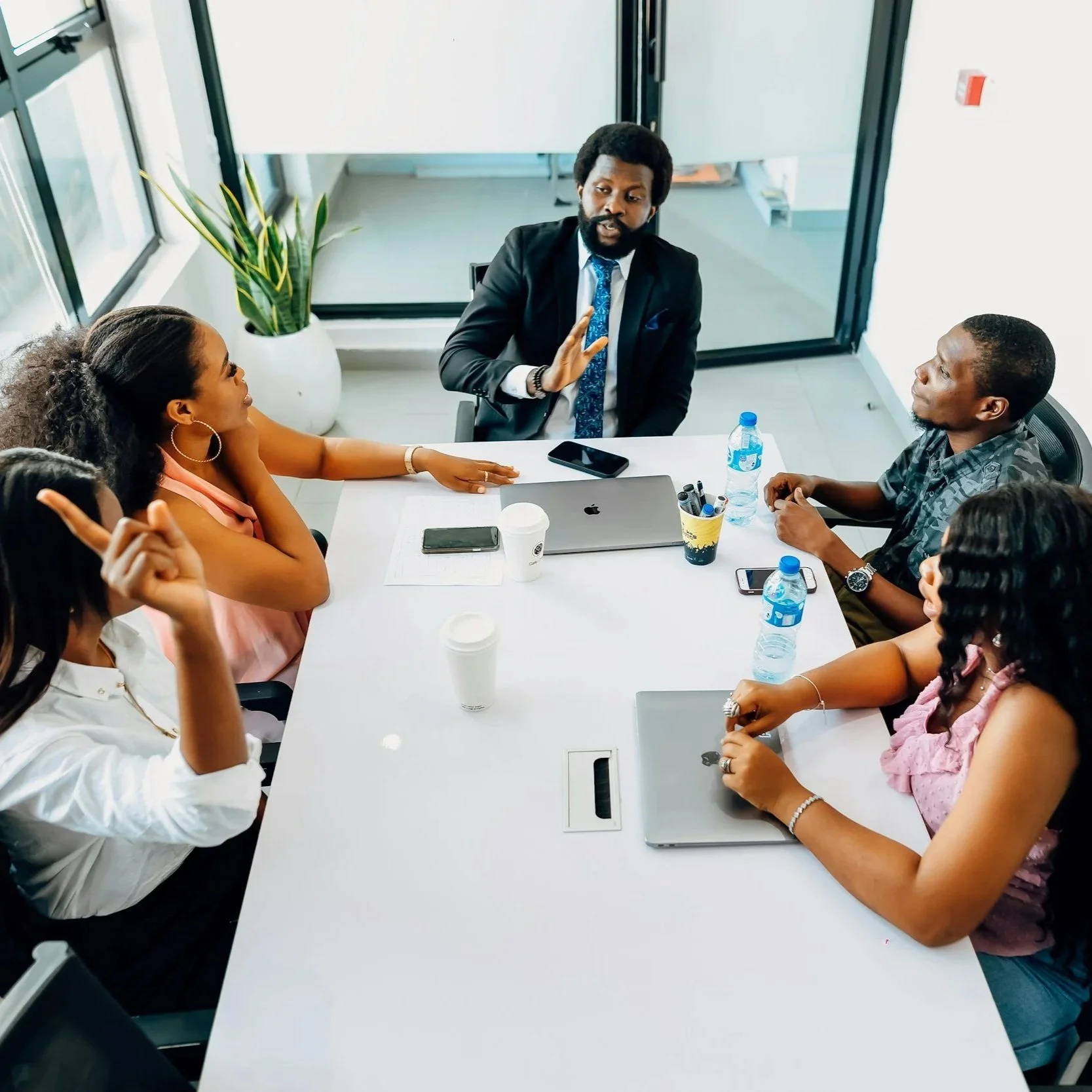 A group of six diverse professionals having a discussion around a white conference table in a modern office. The man in a suit appears to be leading, with others listening attentively. The table has laptops, phones, water bottles, and coffee cups. There is a large pot with a plant, front windows, and a glass door in the background.