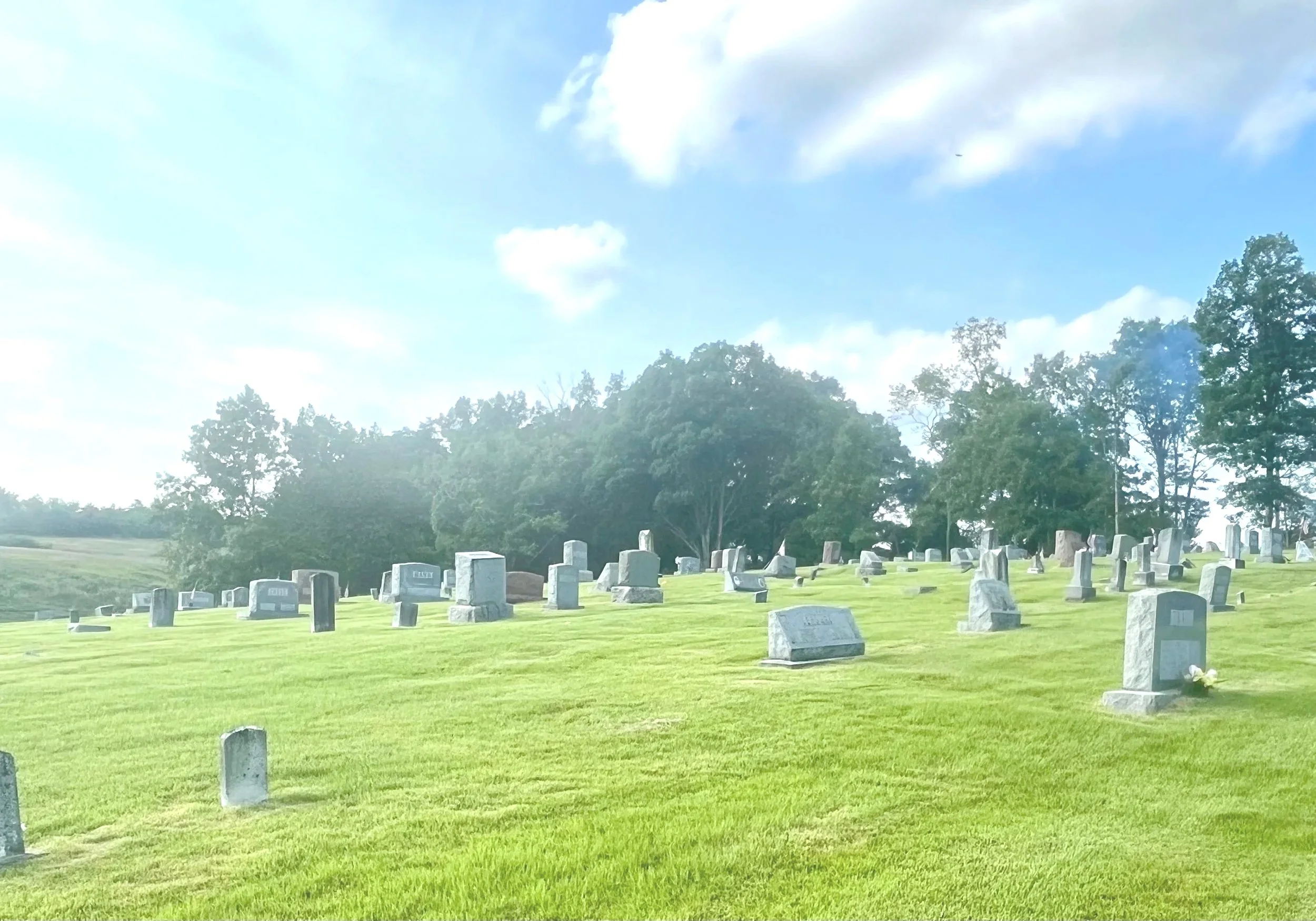 A cemetery with numerous gravestones on a grassy hill, trees in the background, and a partly cloudy sky overhead.