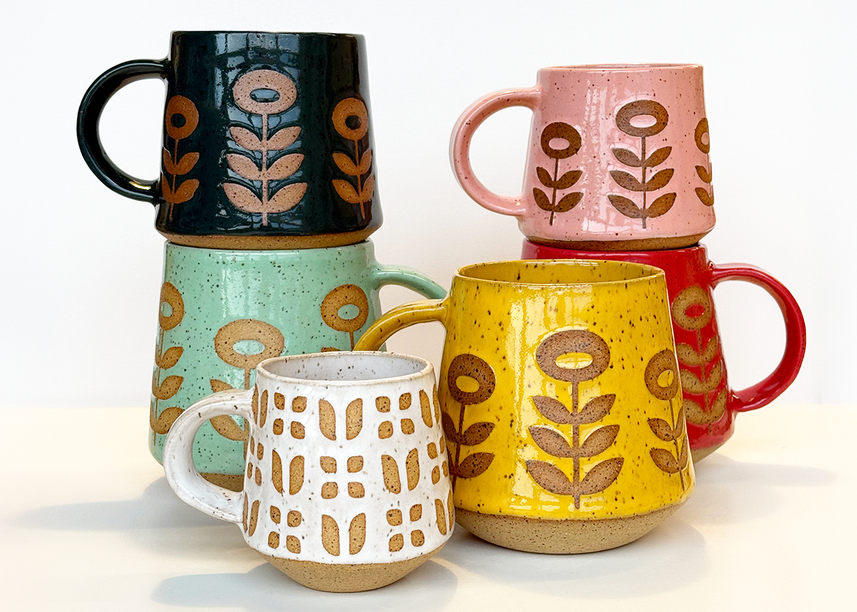 Colorful hand built ceramic mugs with floral mid century modern designs stacked and arranged on a white surface.