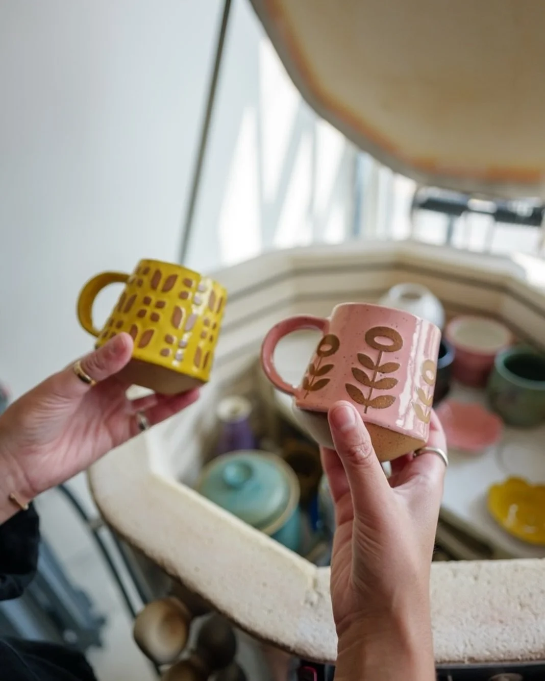 Two hands holding colorful handmade ceramic mugs with decorative patterns, with a pottery wheel and other ceramic items in the background.
