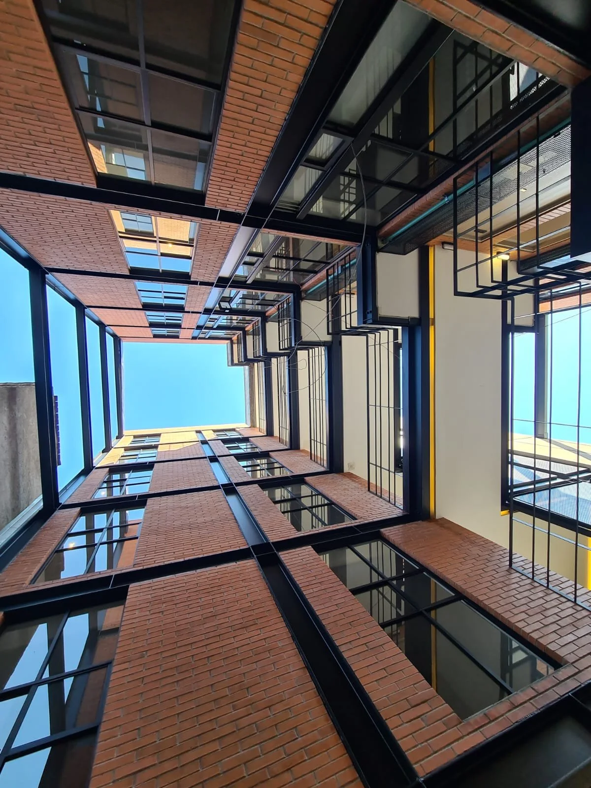 Vista desde abajo de un patio de edificio de varios pisos con paredes de ladrillo, ventanas de vidrio y estructuras metálicas negras y amarillas, hacia un cielo azul.