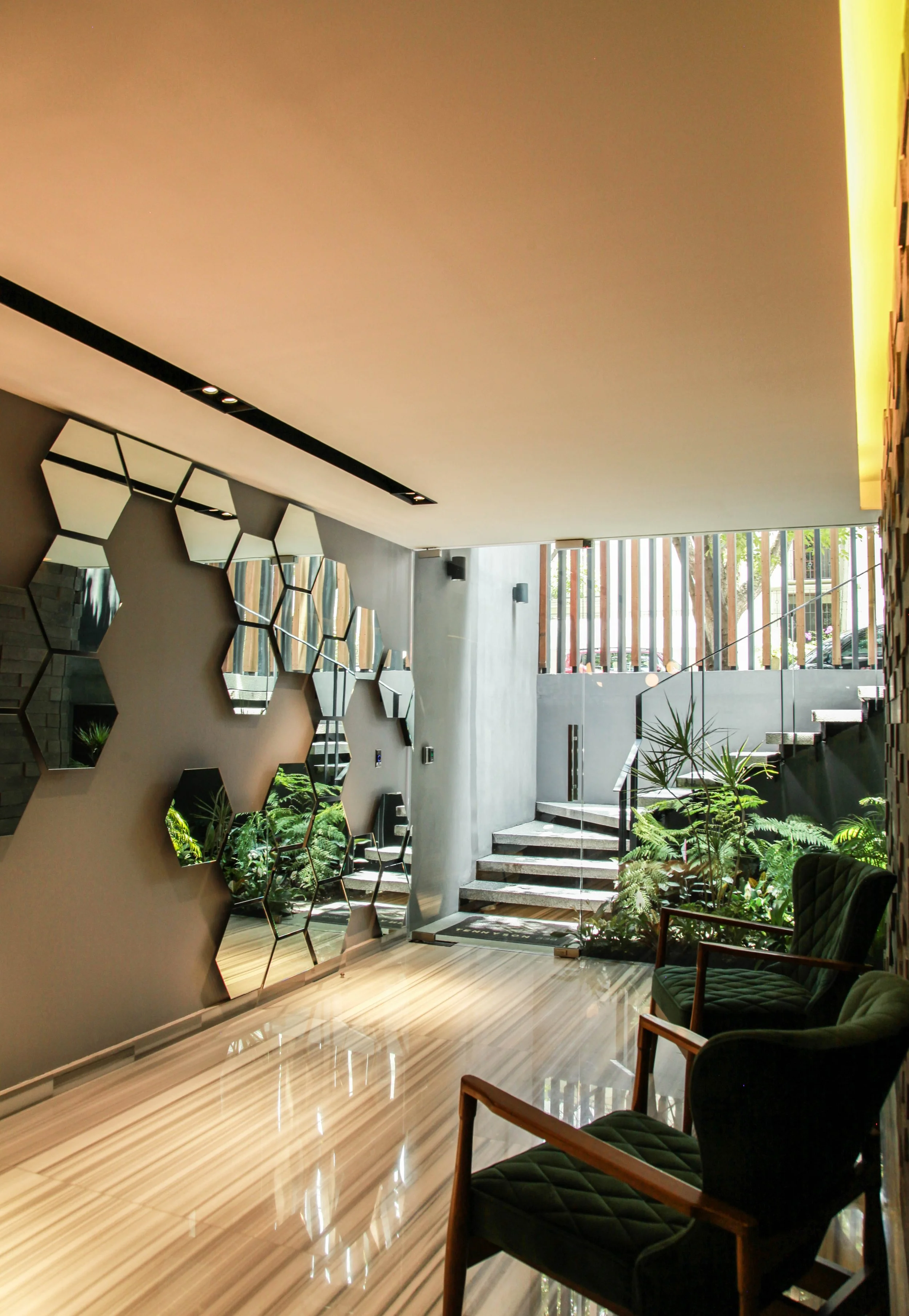 Interior moderno con espejos hexagonales en la pared, escaleras de mármol, plantas, ventanas con barras de madera y sillas tapizadas en verde, con suelo de madera pulida.