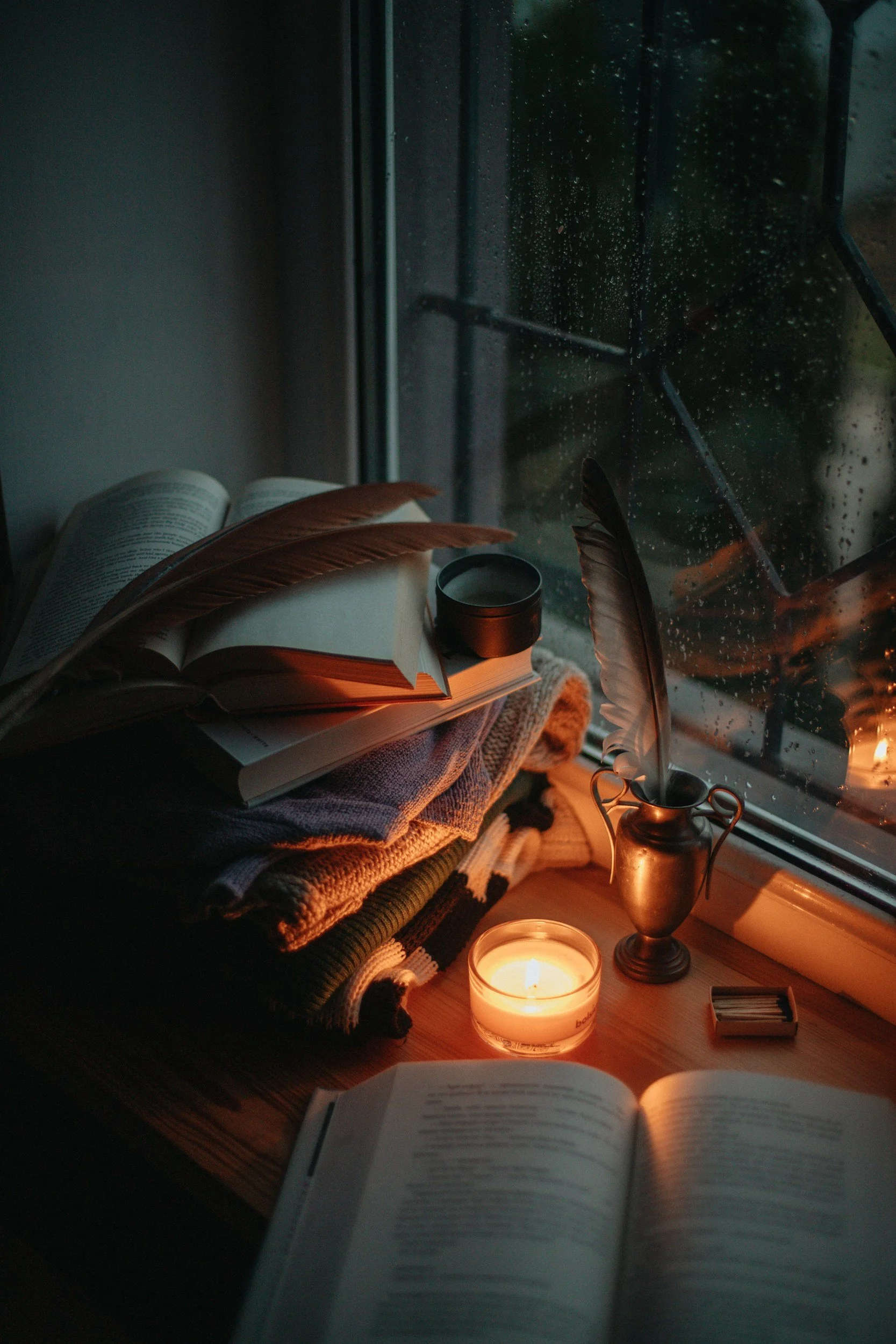A cozy reading nook by the window on a rainy day, with an open book, a lit candle, a decorative pitcher, and a cup, surrounded by folded blankets and a feather quill.