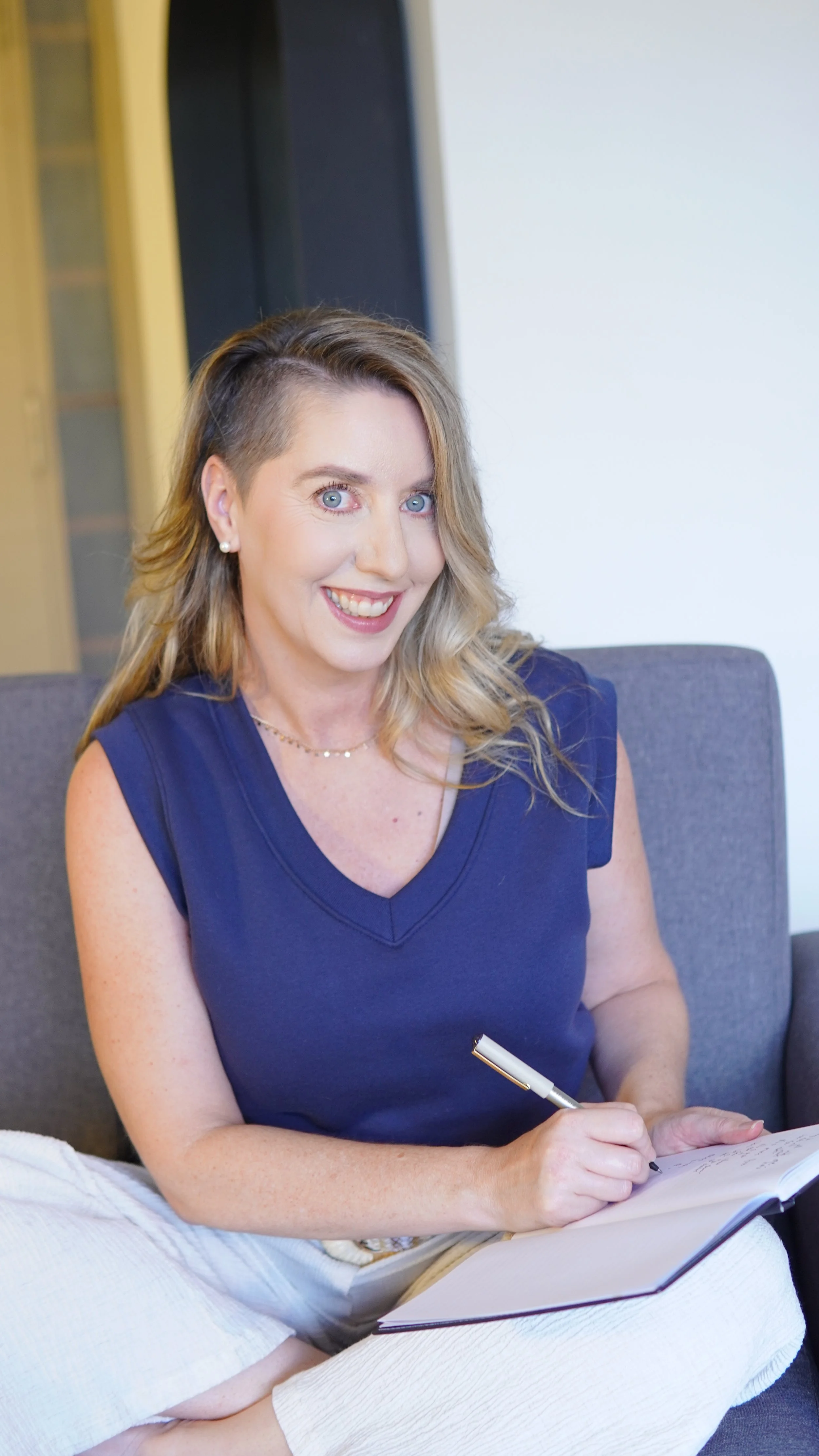 Smiling woman in a navy blue top writing notes in a notebook with a pen.