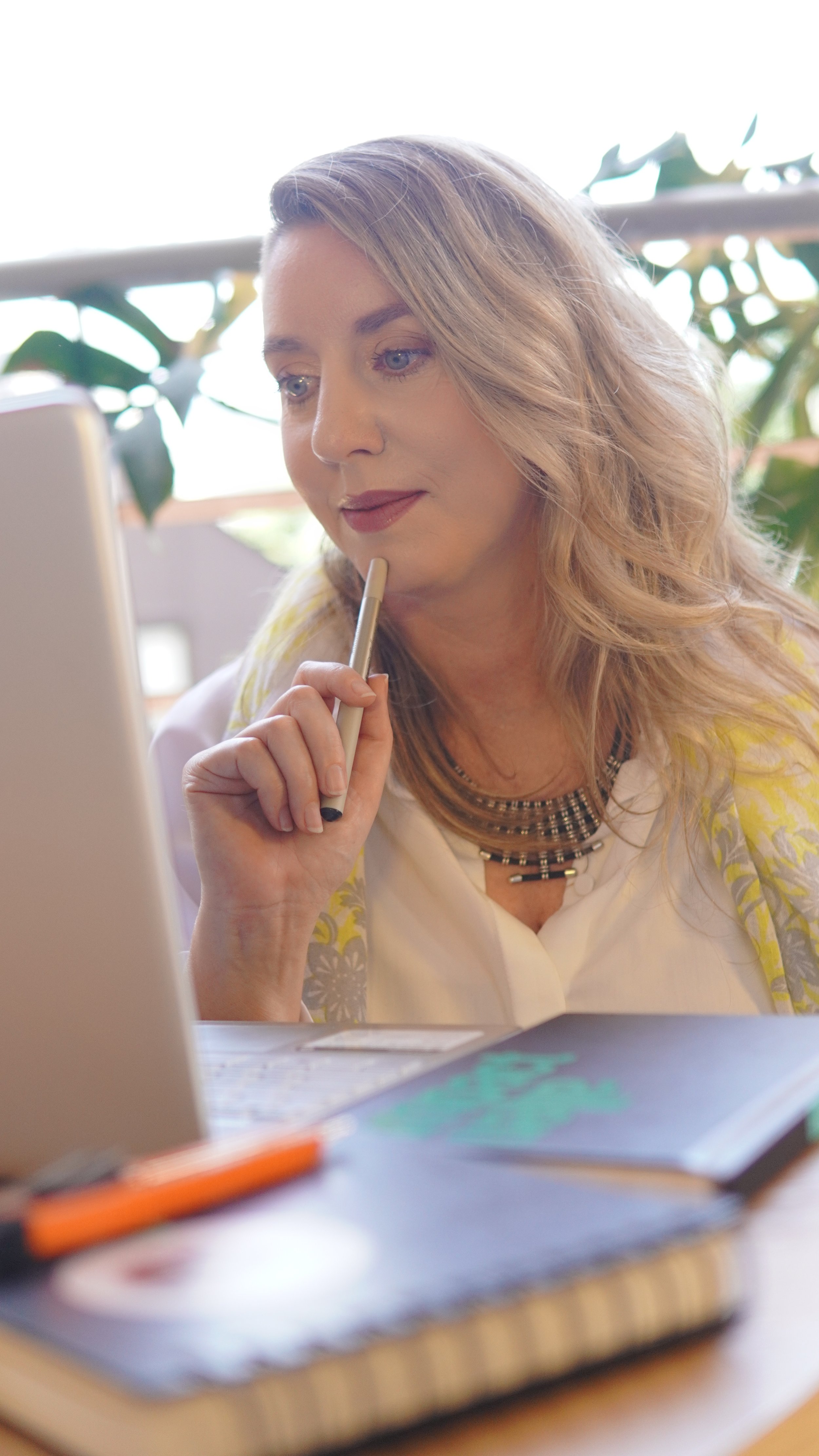 Close-up of a blonde woman holding a pen near her lips while looking at a laptop.