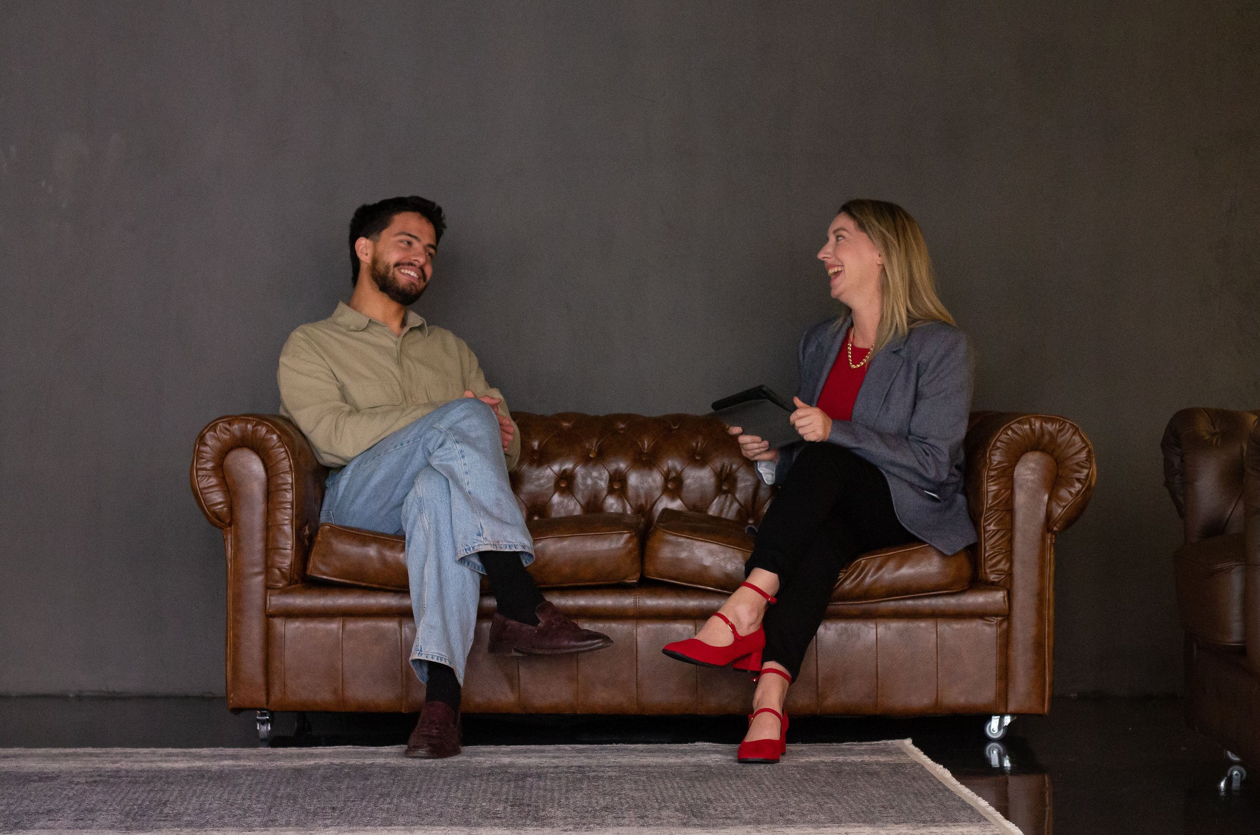 Two people laughing and talking while seated on a brown leather Chesterfield sofa.