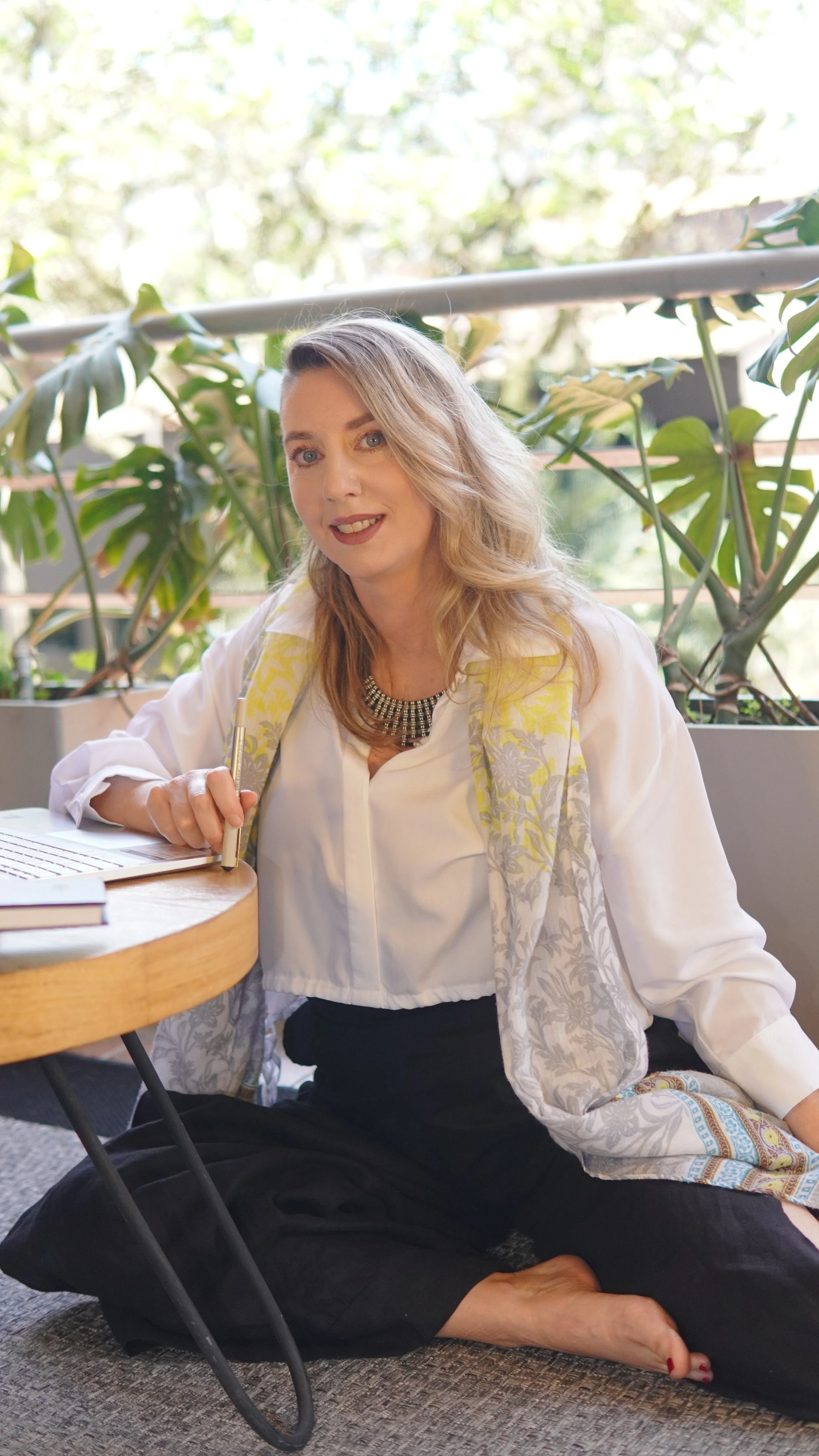 Smiling woman in a white blouse and patterned scarf writing at a table surrounded by green plants.