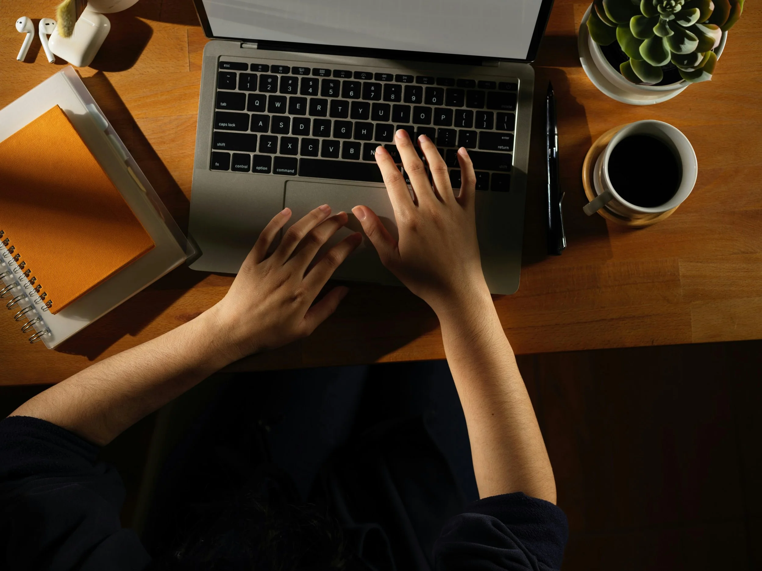 Overhead view of a person working on a laptop at a wooden desk, with a potted succulent plant, a cup of coffee, a pen, a notebook, and wireless earbuds on the desk.