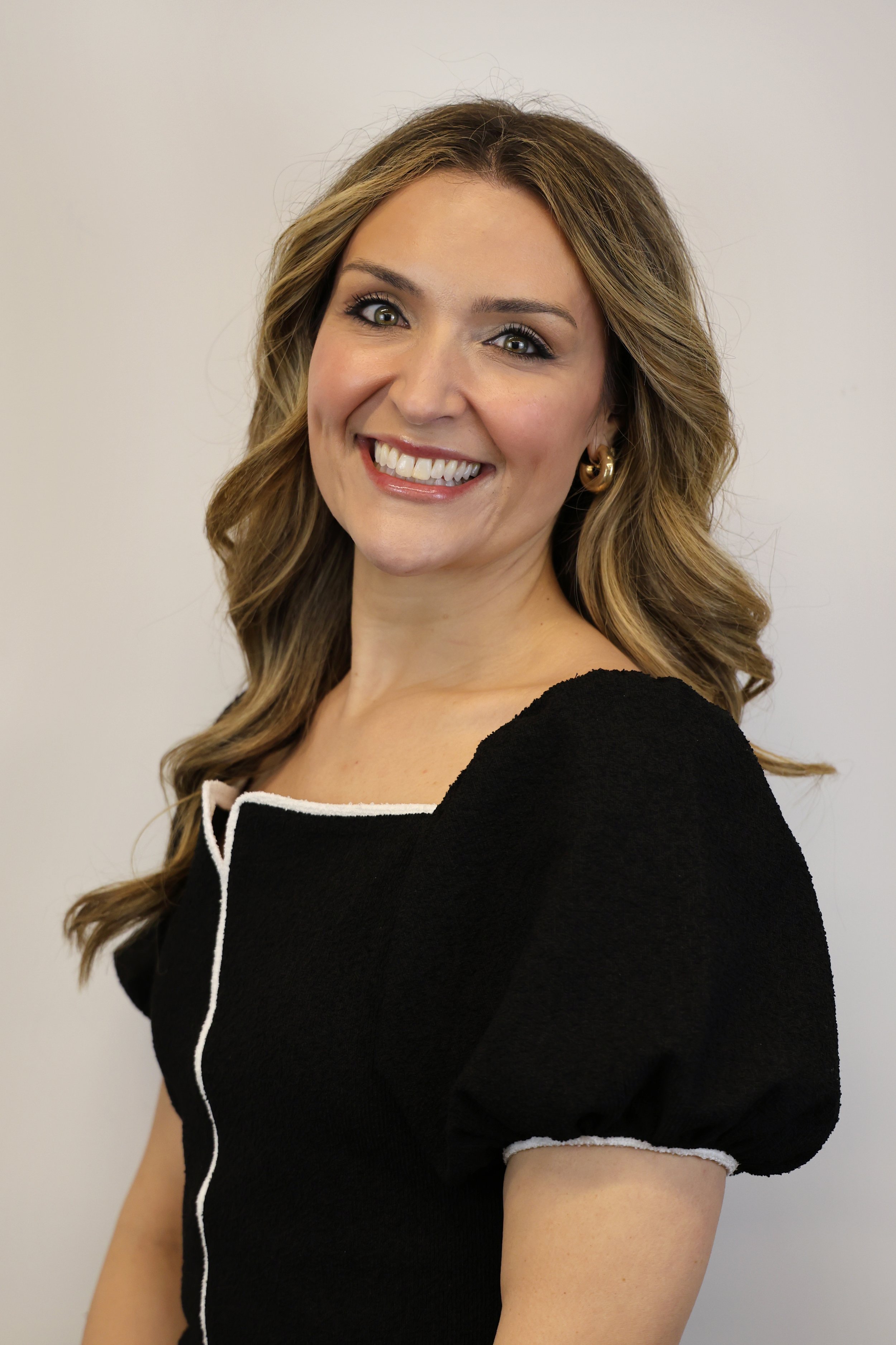 A smiling woman with blonde wavy hair wearing a black top with white trim and gold hoop earrings standing against a plain white wall.