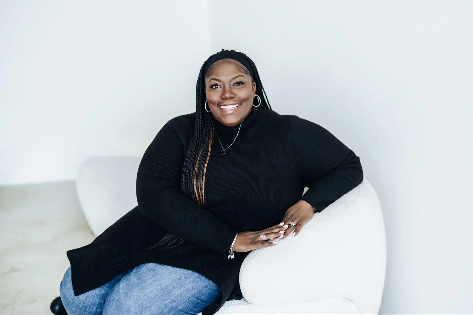 A woman with dark hair in braids, wearing a black top and silver jewelry, sitting on a white chair and smiling.