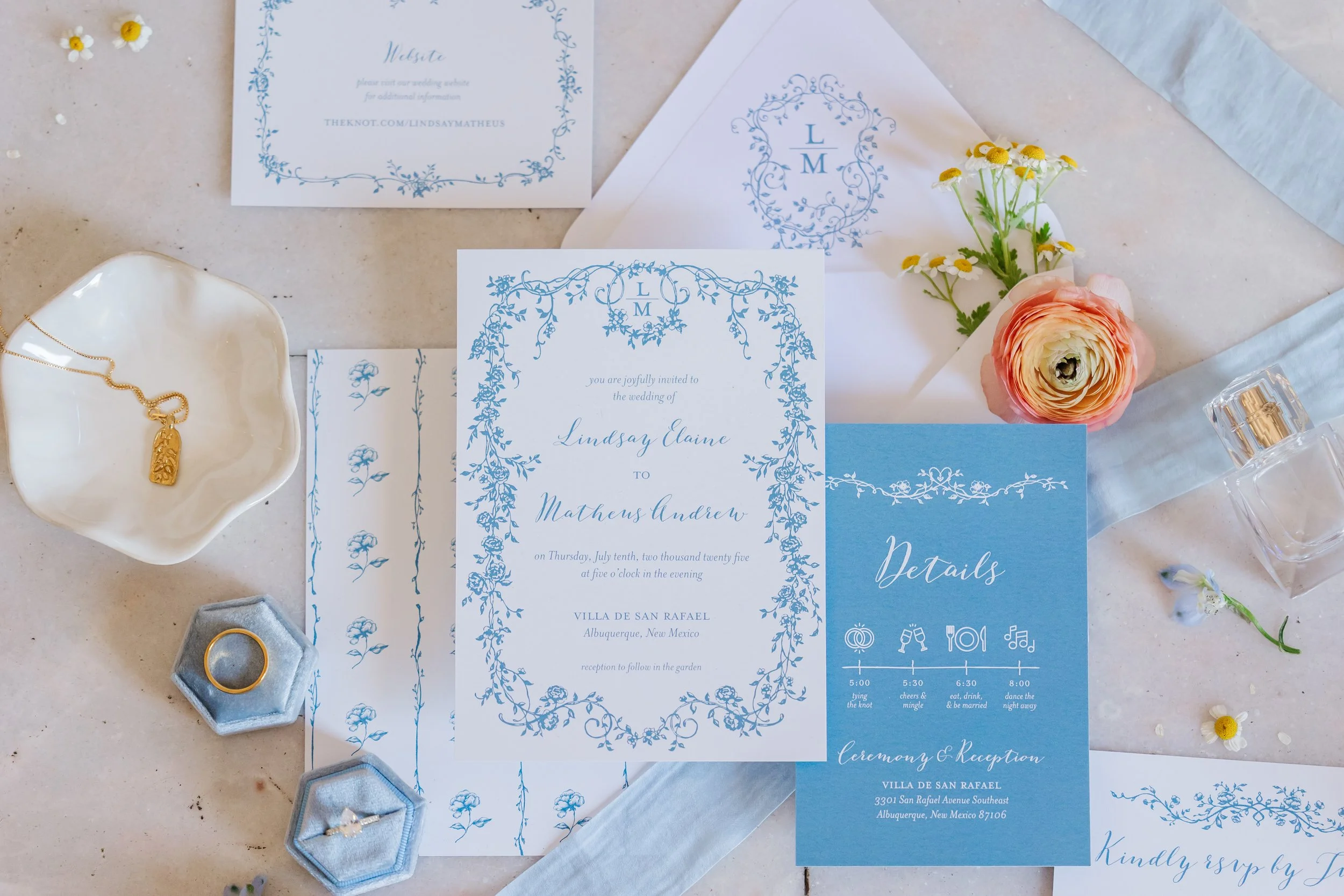 Wedding invitation, details card, and RSVP card with blue floral designs, gold ring and necklace, flower, perfume, and floral decoration on table.