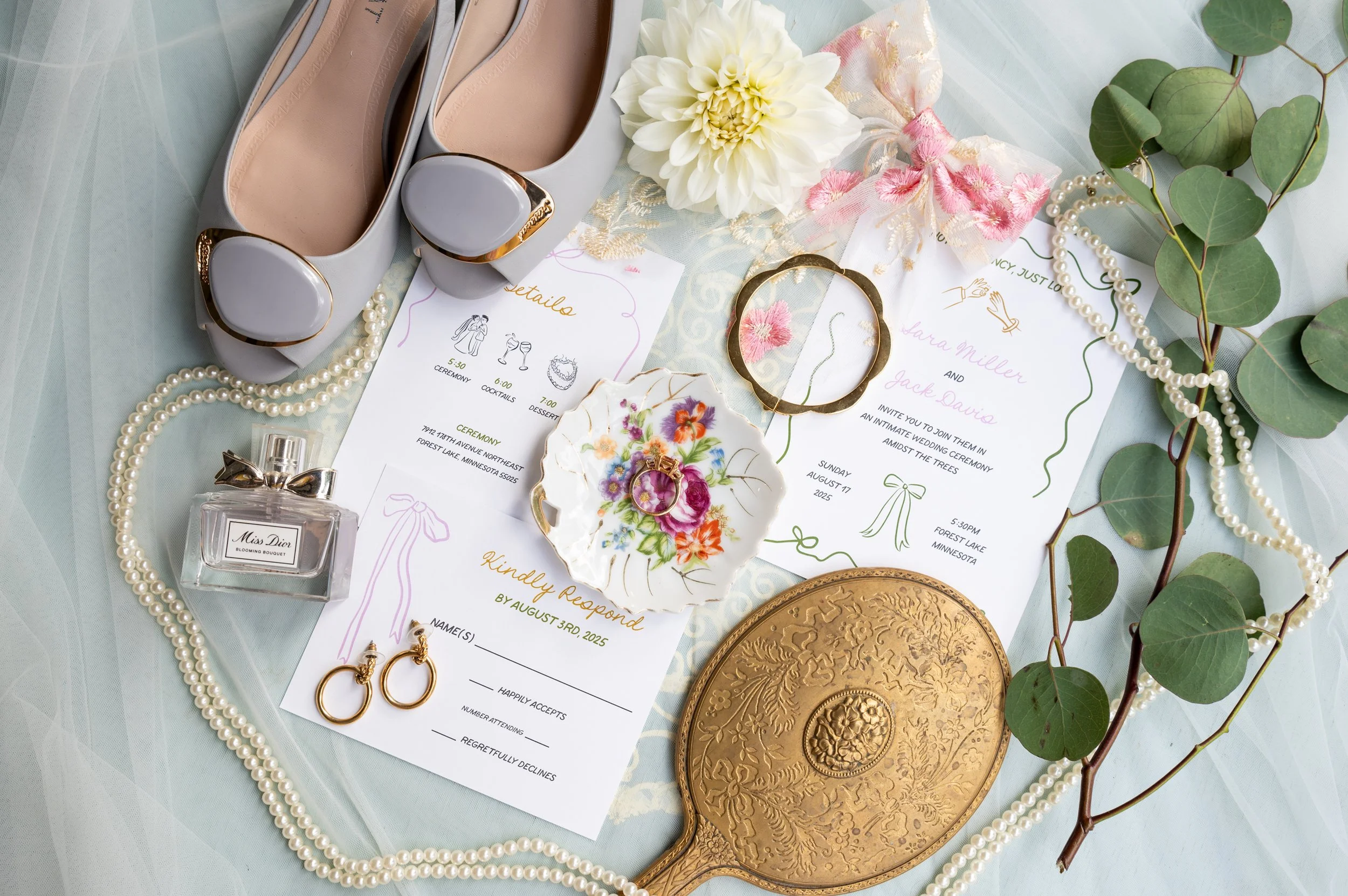 Wedding invitation, shoes, pearl necklace, earrings, perfume, and decorative items arranged on a cloth, with flowers and greenery, preparing for a wedding.