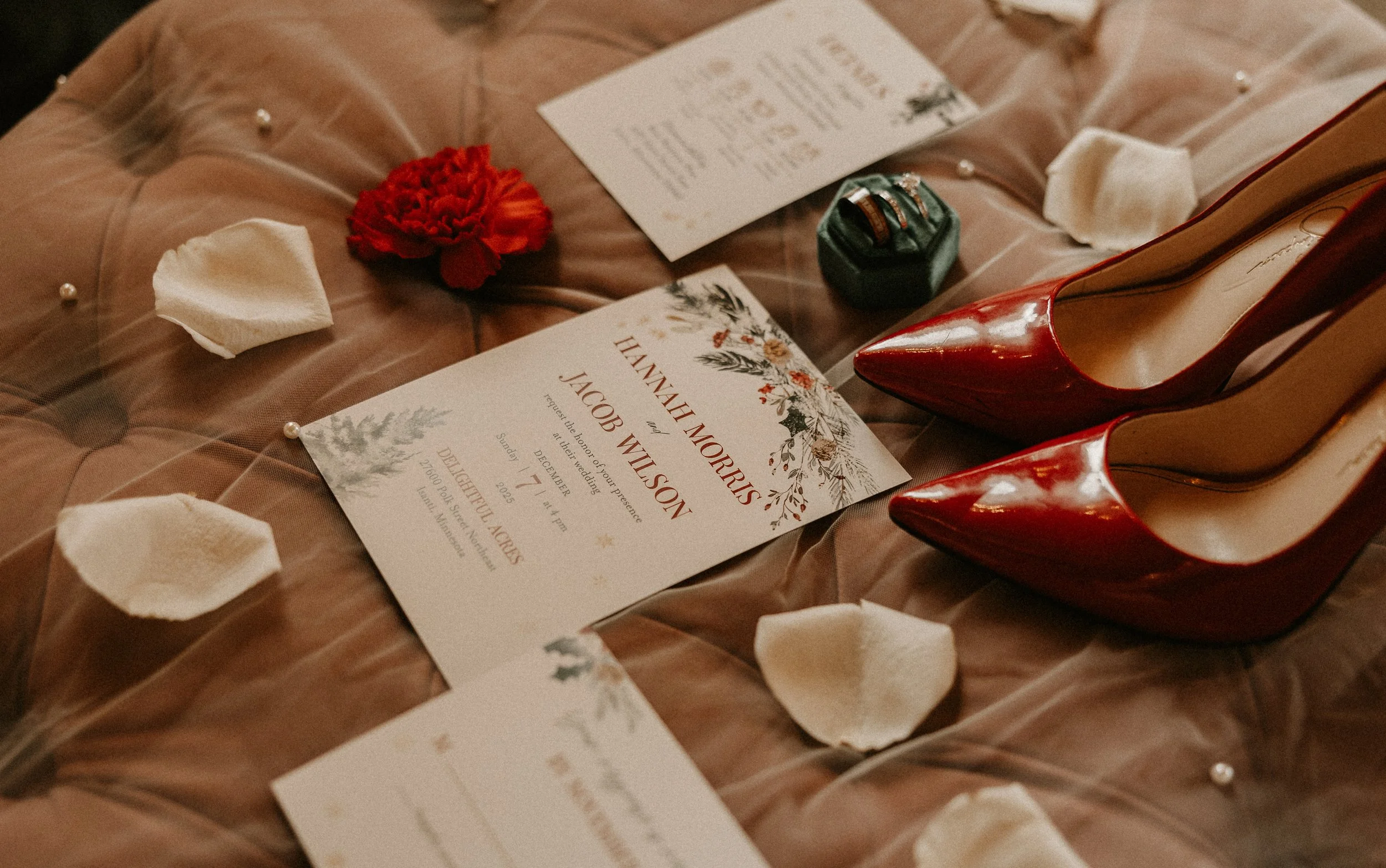 Wedding invitation, red high heels, red flower, pearl accents, ring box with rings, and scattered white petals on a satin fabric background.