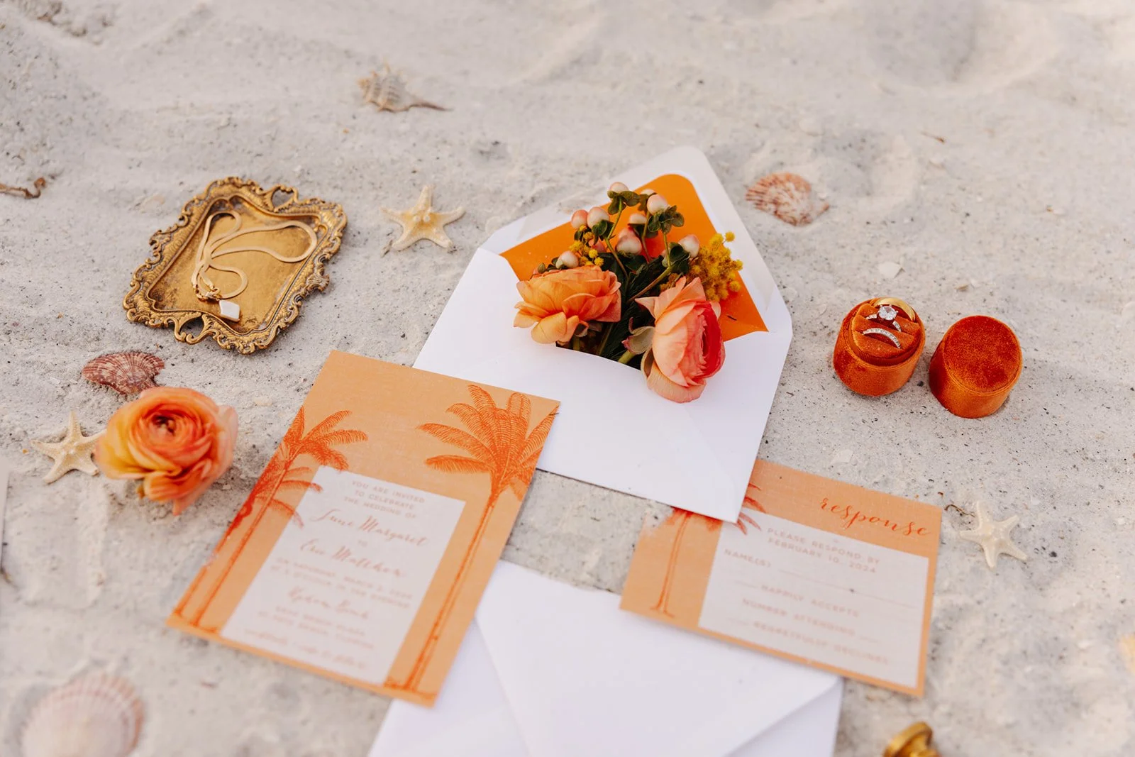 Beach wedding invitation with coral-colored flowers, seashells, starfish, jewelry, and orange response card on sandy surface.