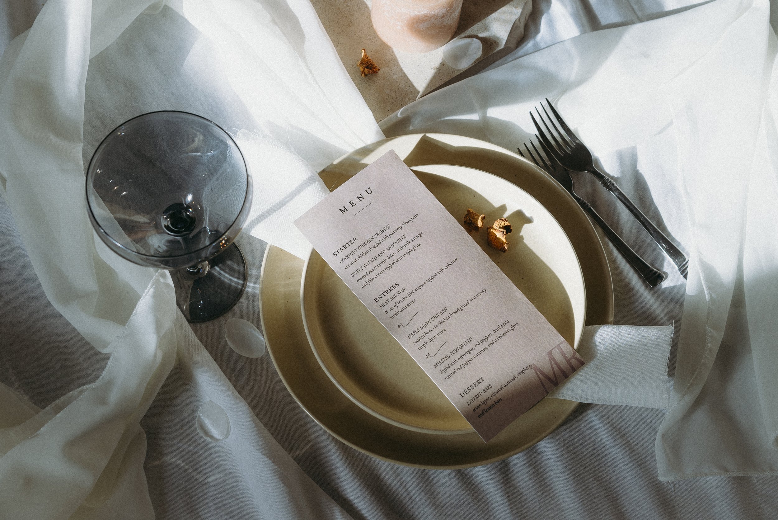 A dining table setting with an empty wine glass, stacked beige plates with a printed menu on top, silverware, and some small decorative food items on a linen tablecloth with sheer white fabric draped around.