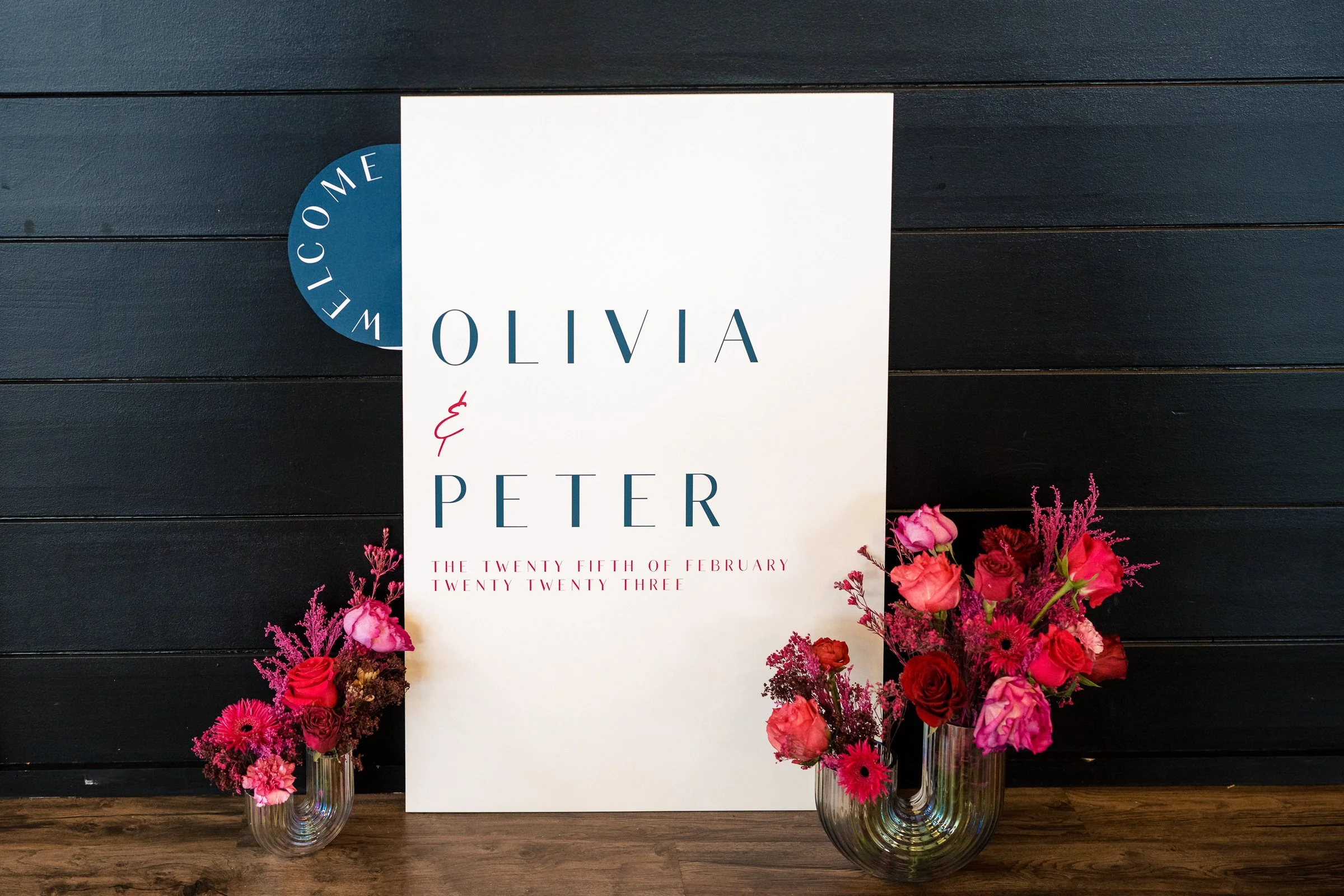 Wedding welcome sign with pink and red flowers in two vases, black wooden background.