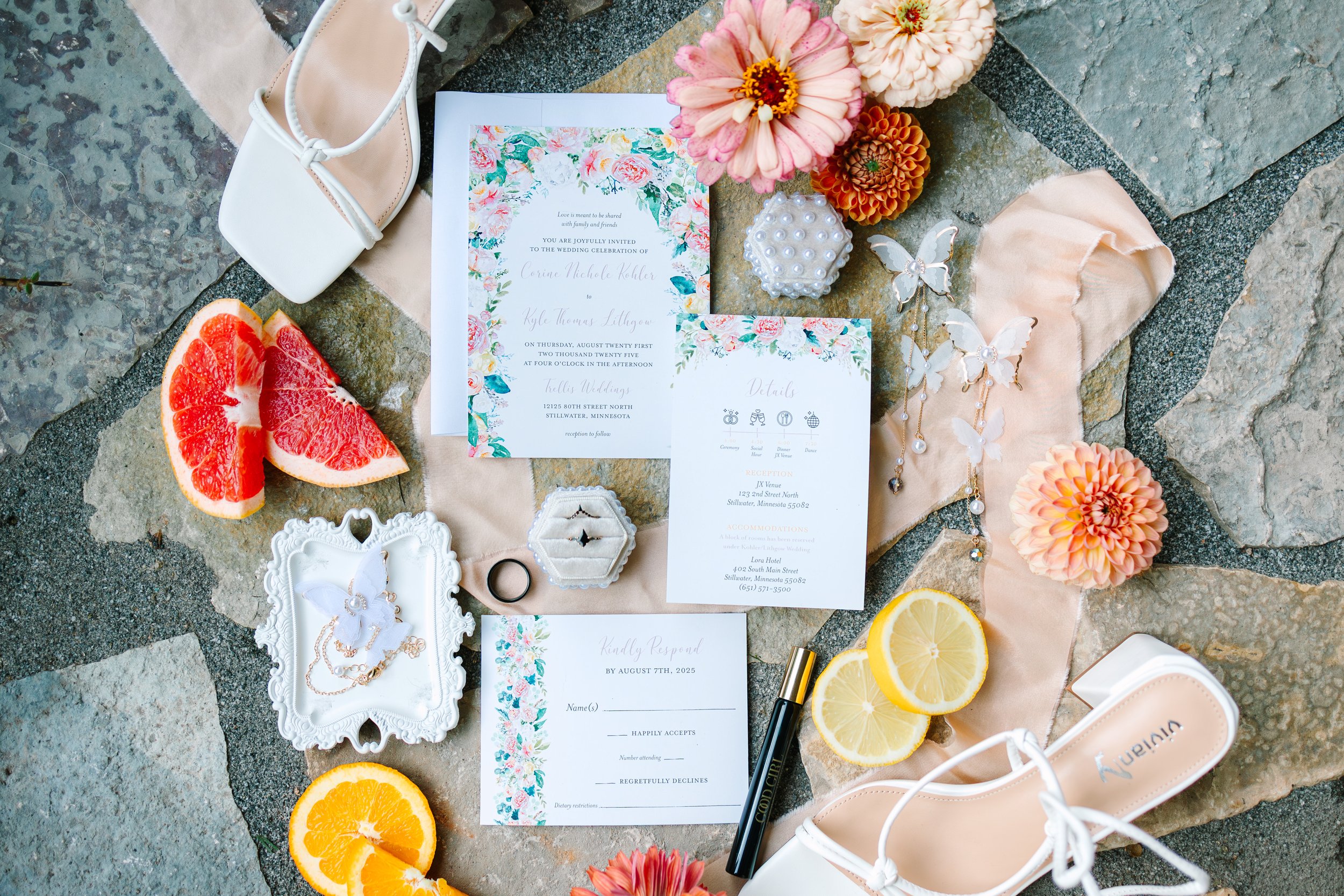 Wedding invitation suite with floral borders, surrounded by pink and orange flowers, sliced lemons, a grapefruit wedge, white shoes, jewelry, and decorative butterflies on a stone surface.