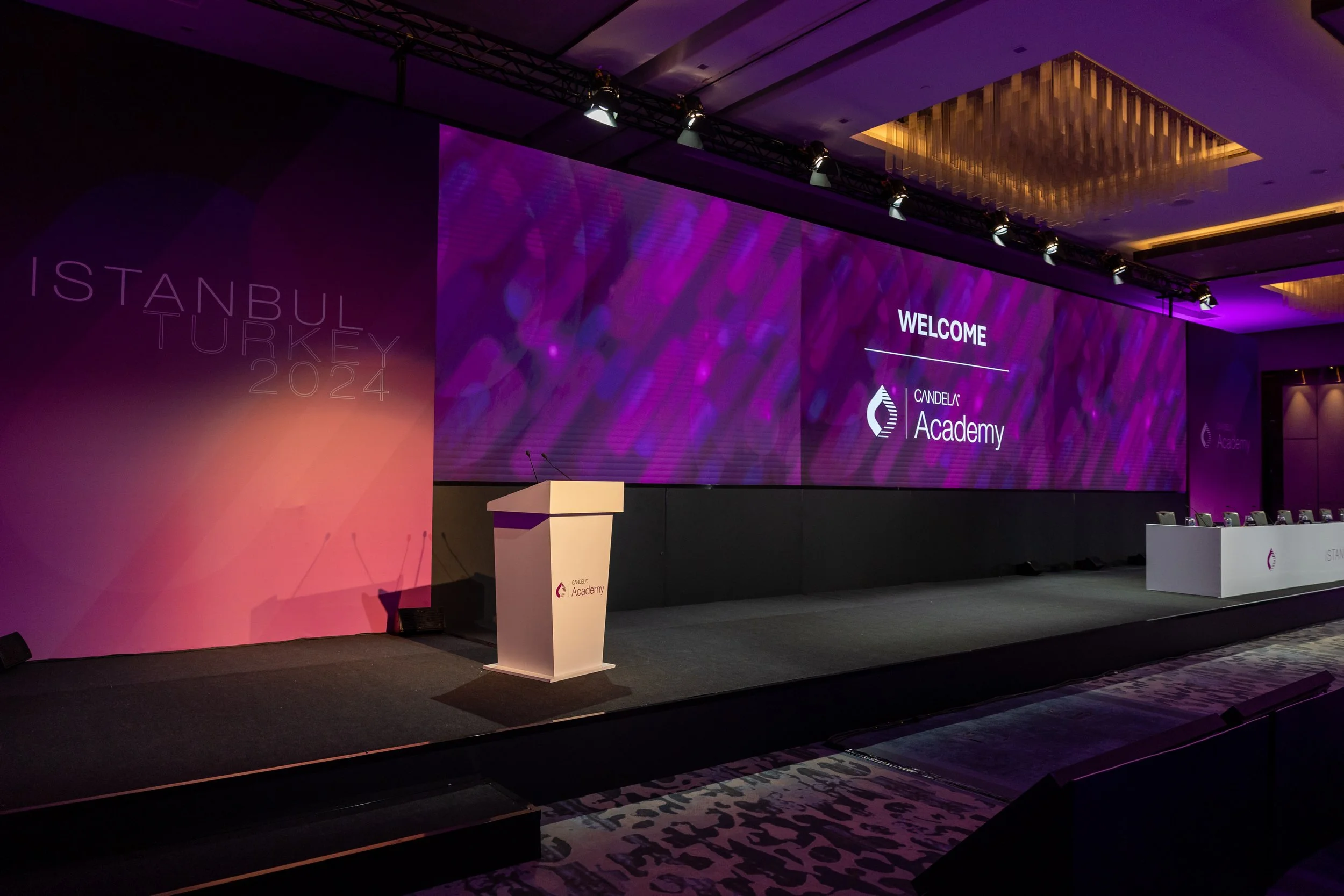 Conference room stage with large screen displaying 'Welcome' and 'Candela Academy' logo, purple lighting, podium at front, chairs along the side, and decorative ceiling lighting.
