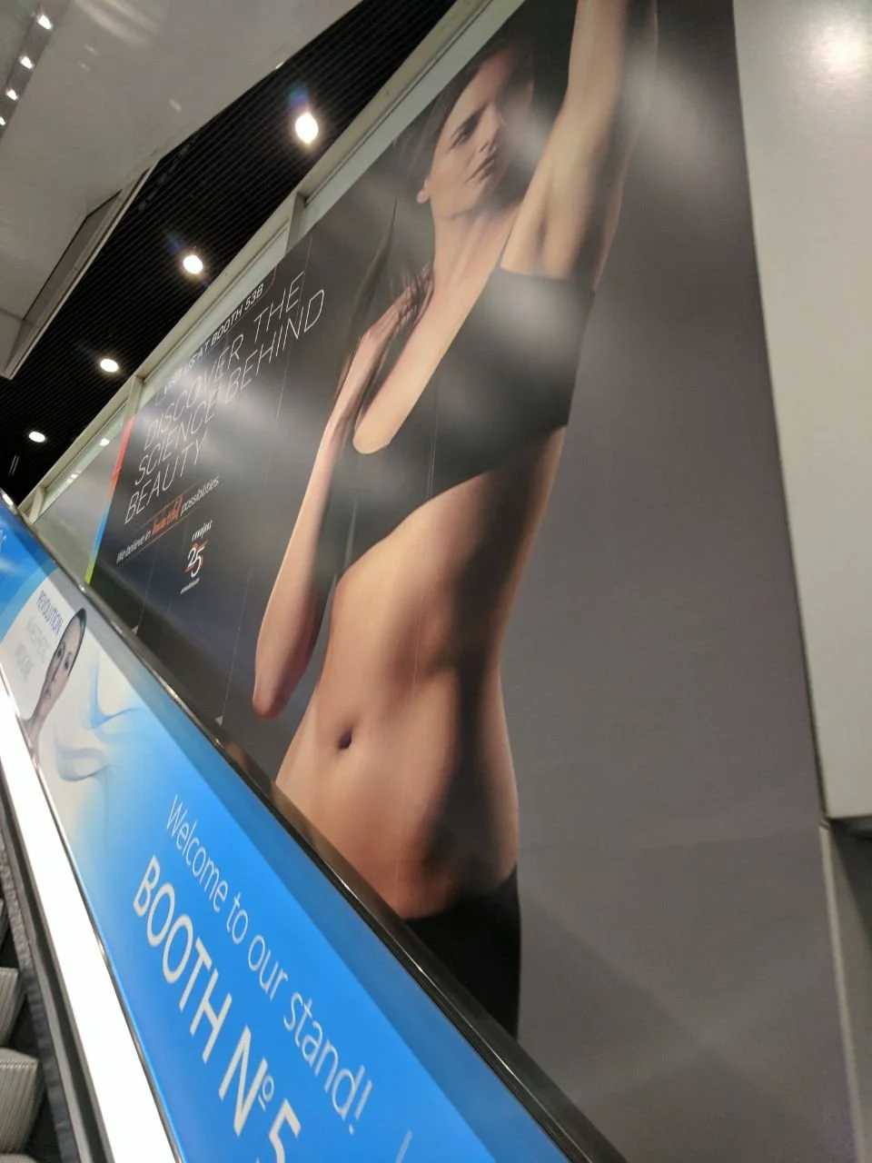 Large advertisement featuring a woman in a black sports bra and shorts with her arms raised, promoting health and beauty. Additional signage includes welcoming text for a booth.