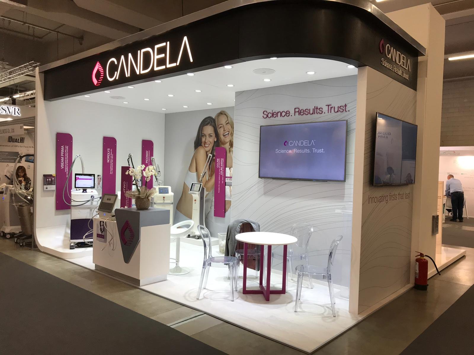 Booth display for Candela at a trade show. It features medical or aesthetic devices, a seating area with clear chairs and a small table, promotional posters with smiling women, and digital screens displaying the brand and tagline 'Science. Results. T