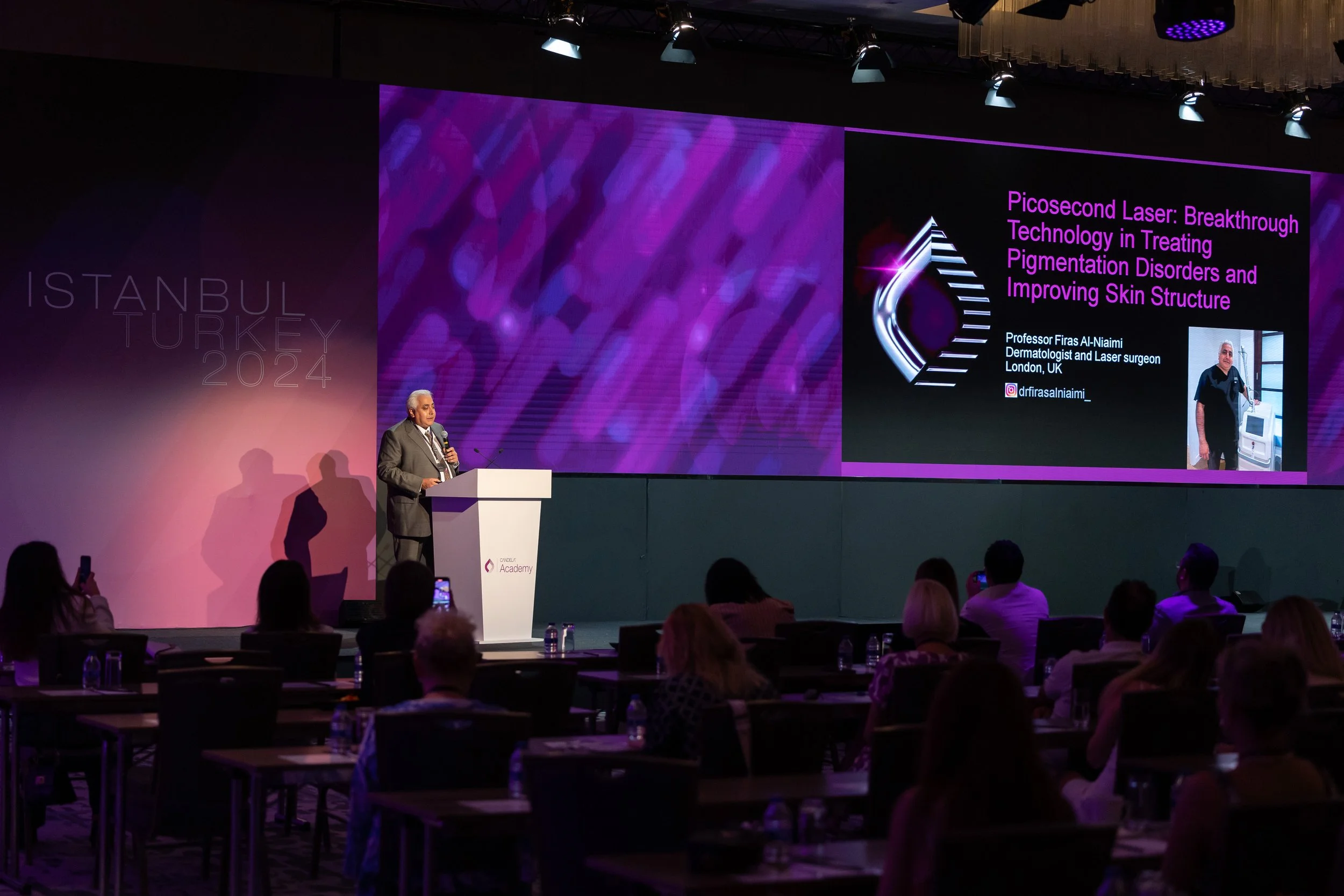 A man in a suit giving a presentation on stage at a conference, with a large screen displaying a slide about pico second laser technology and a photo of the presenter. Audience members are seated in front of him, some taking photos.