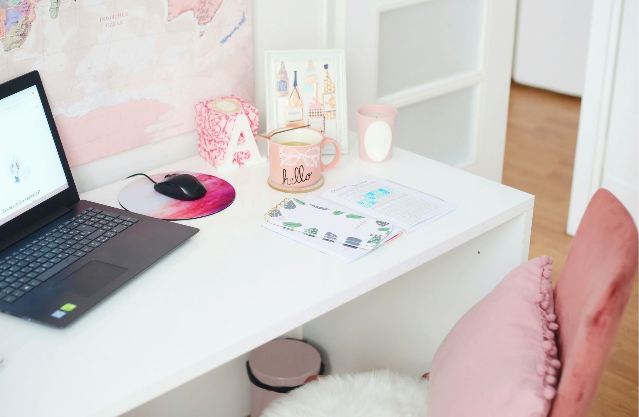A white desk with a laptop, a computer mouse on a pink and purple mouse pad, three pink cups with white designs, and various papers, including a planner and a calendar. In the background, there's a framed picture, a pink decorative box, and a pink hand-shaped paperweight. A world map with pink and pastel colors is on the wall behind, and a person's arm in a pink, fluffy sleeve is partially visible at the bottom right corner.