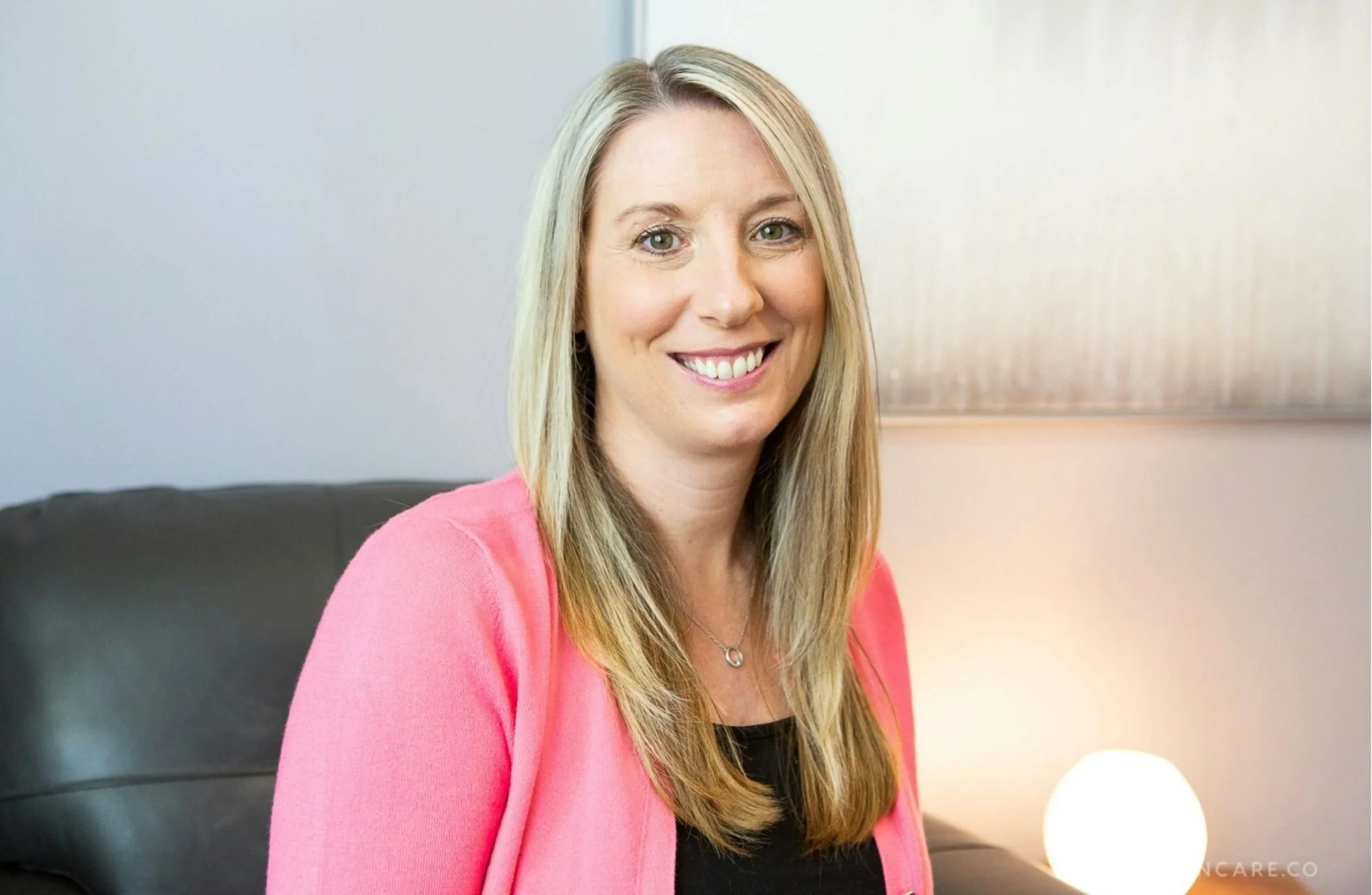 Cadence Chiasson, a certified EMDR therapist in Morrison, CO, with long blonde hair smiling at the camera, wearing a pink cardigan over a black top, sitting on a black sofa with a white spherical lamp beside her.