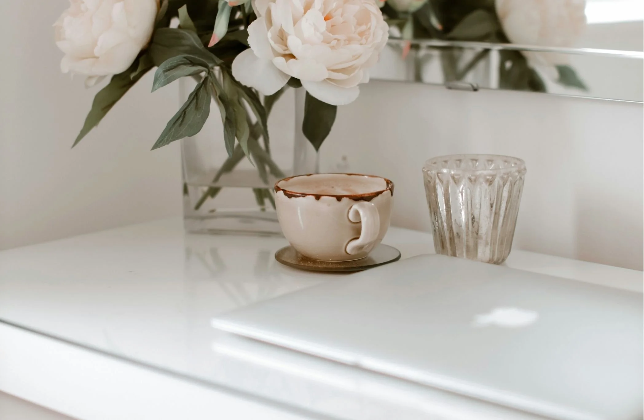 A white shelf with a pink flower bouquet in a clear glass vase, a beige ceramic teacup with a brown rim and a matching saucer, and a clear textured glass cup.