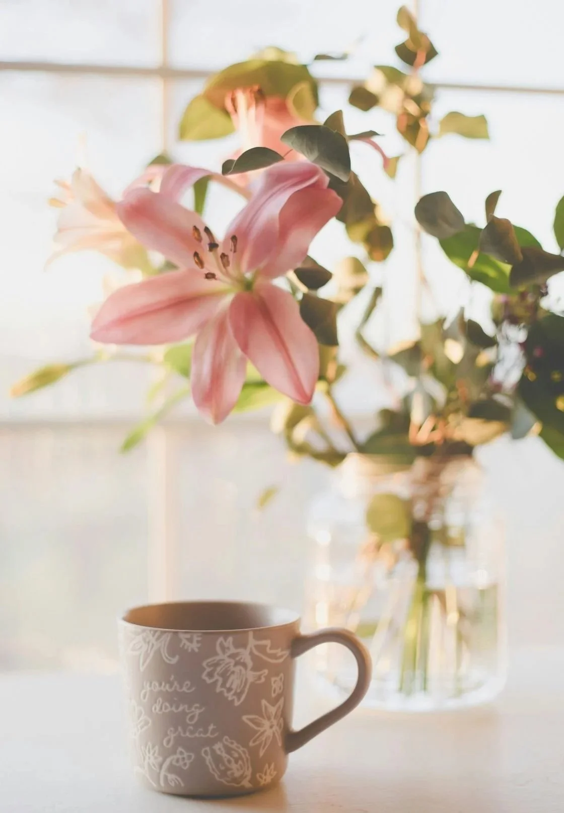 A pink lily flower in a vase on a table, with a coffee mug in the foreground and a window with sunlight in the background.