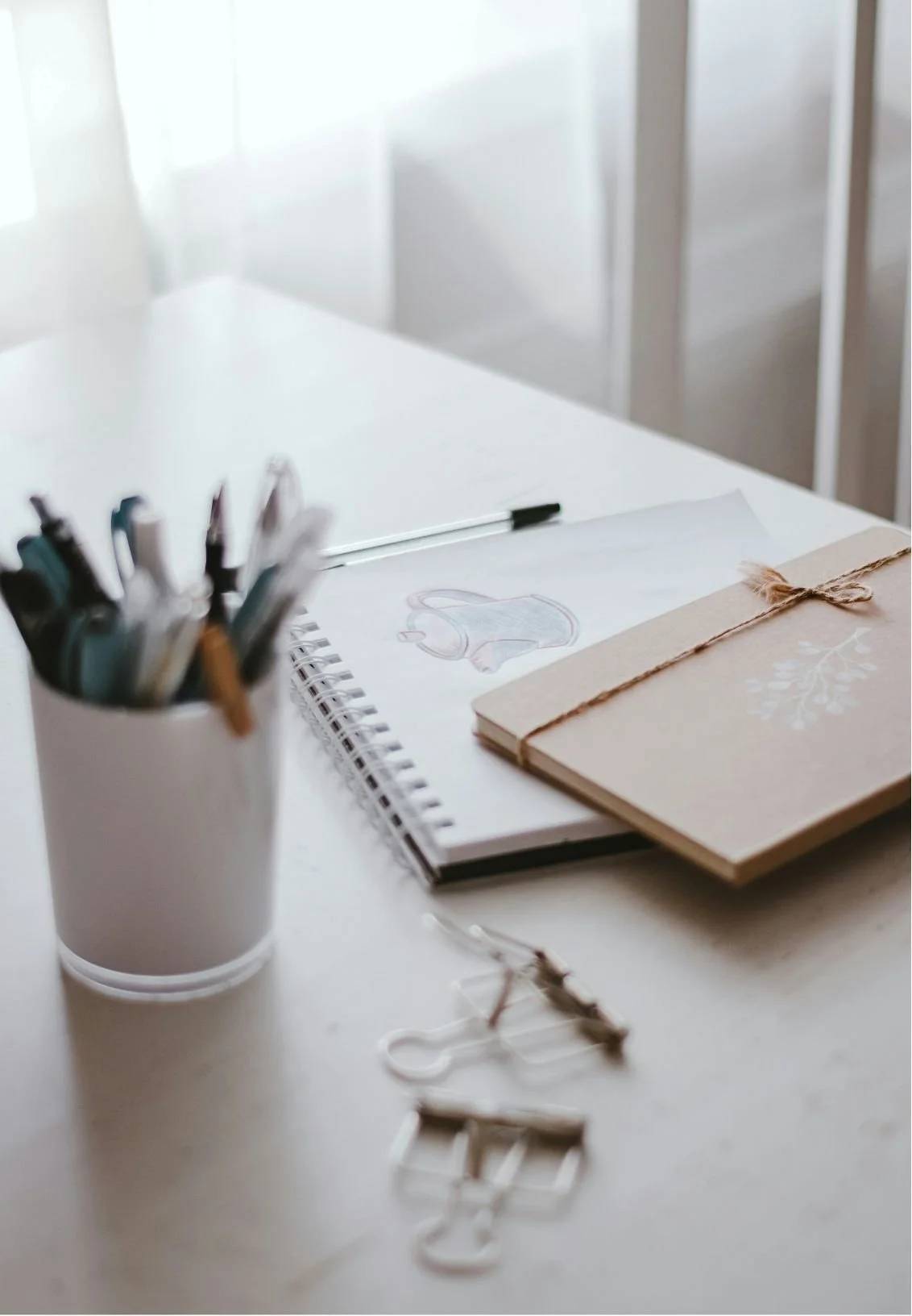 A white desk with a cup of pens, a sketchbook, two notebooks, and paper clips.