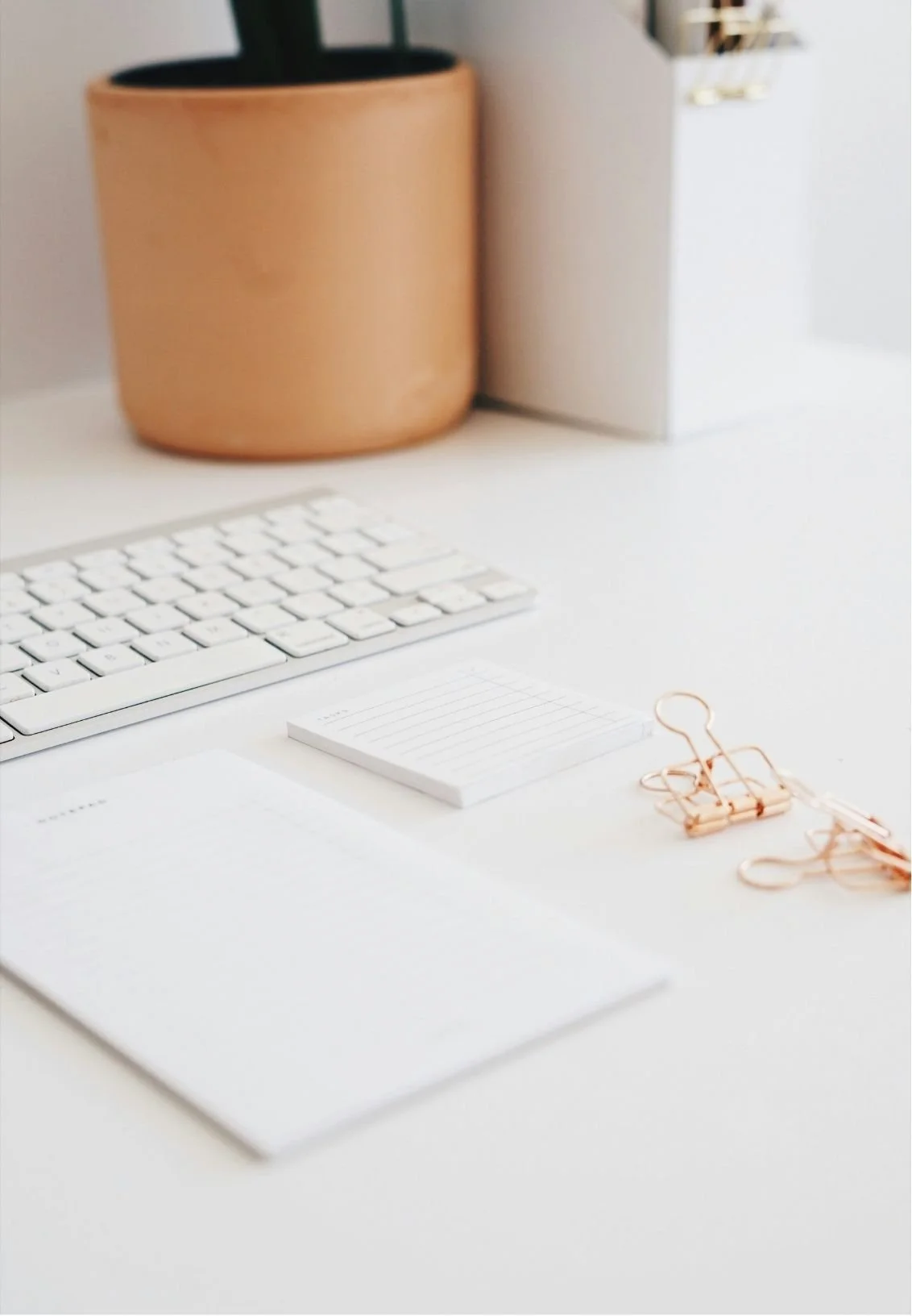A white desk with a computer keyboard, notepad, and gold paper clips. In the background, a potted plant and a file organizer are visible.