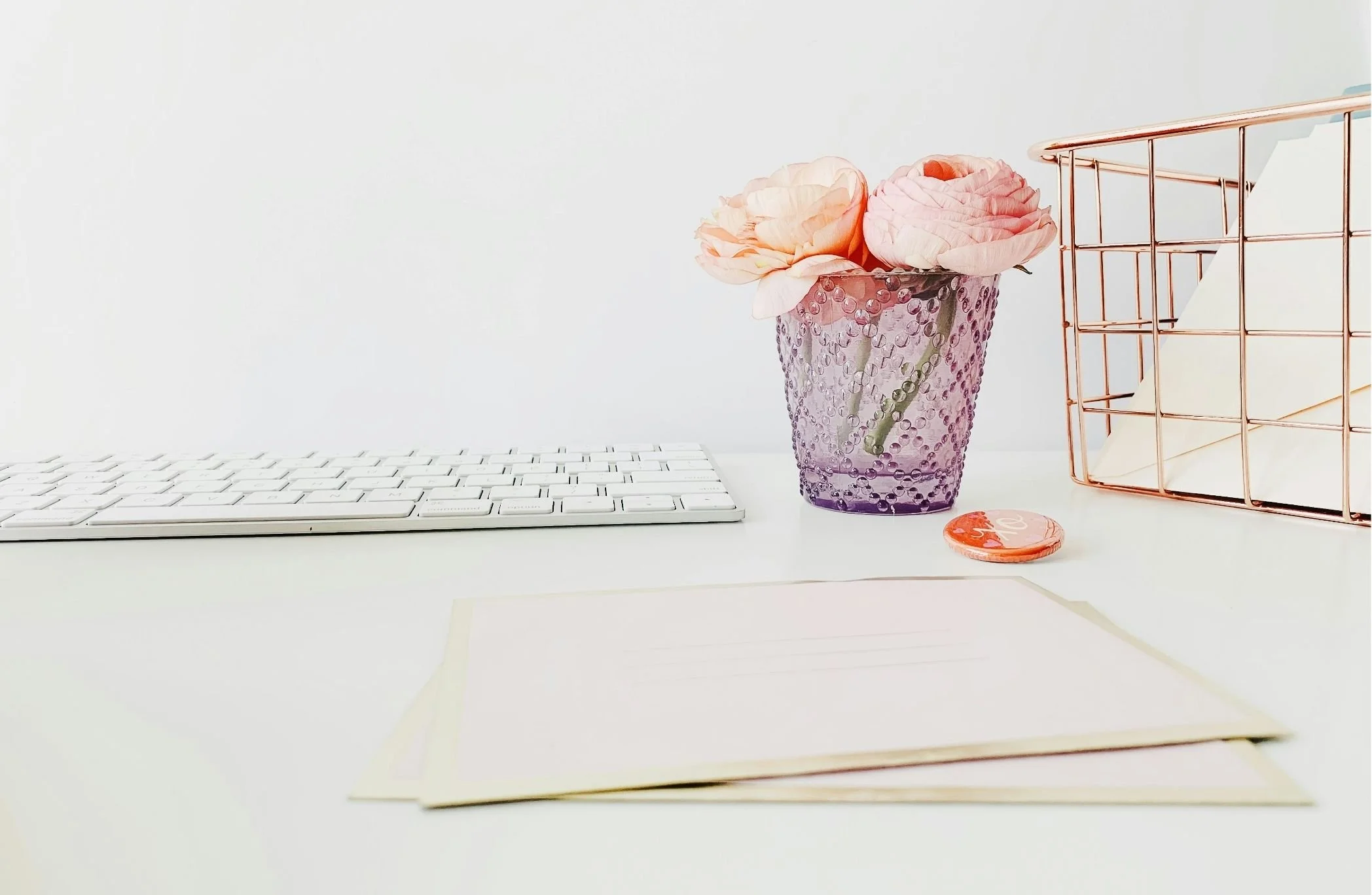 A white desk with a keyboard, a pink glass vase with peach-colored roses, a copper wire file organizer, and some papers and a small orange item. The background is plain white.