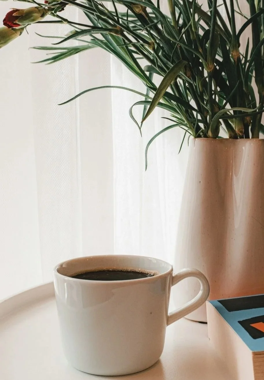 A white ceramic mug filled with black coffee sitting on a white surface, next to a beige vase with green leafy plant, in front of a white curtain.
