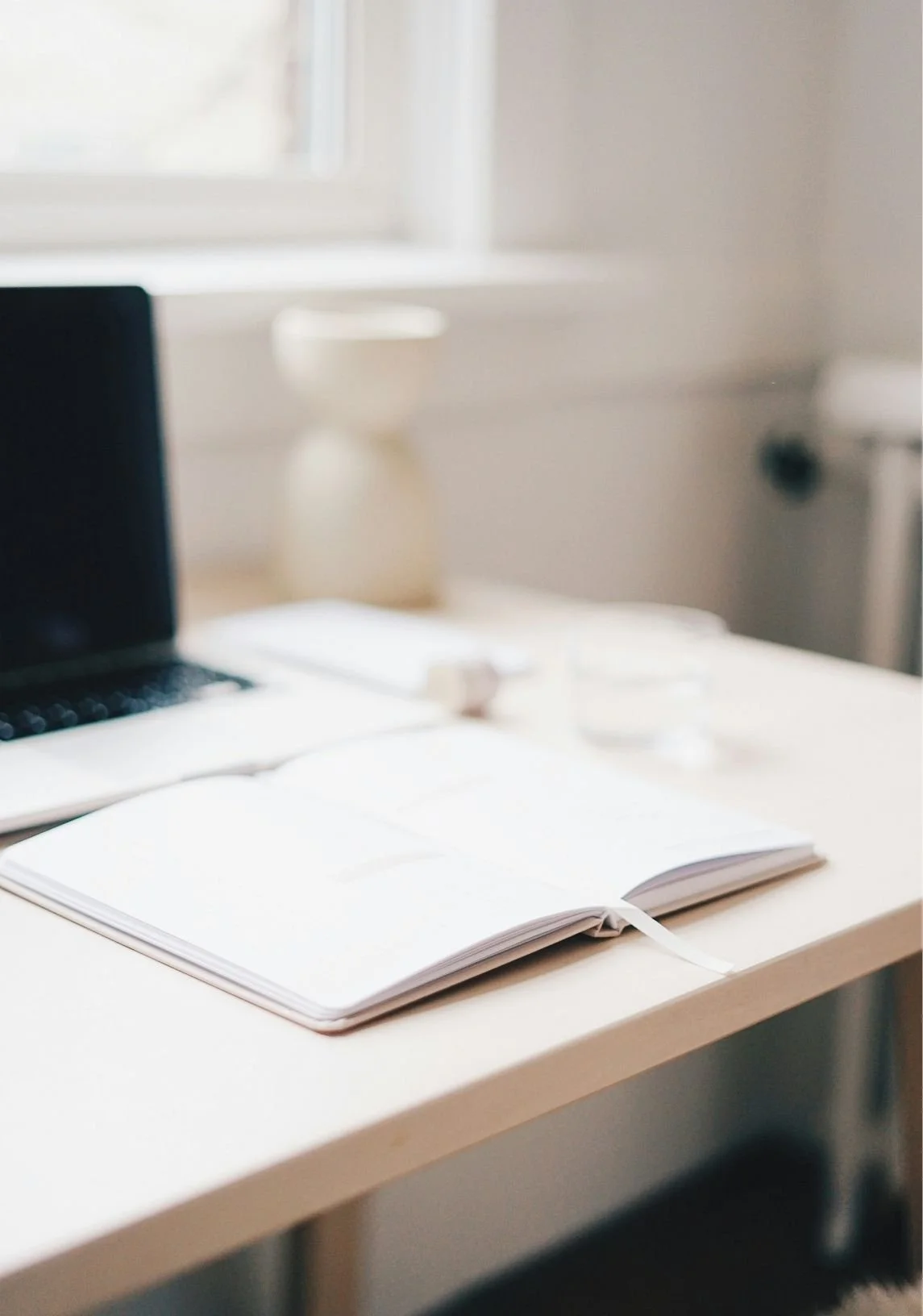 Open notebook on a white desk with a laptop, glass of water, and blurred objects in the background.